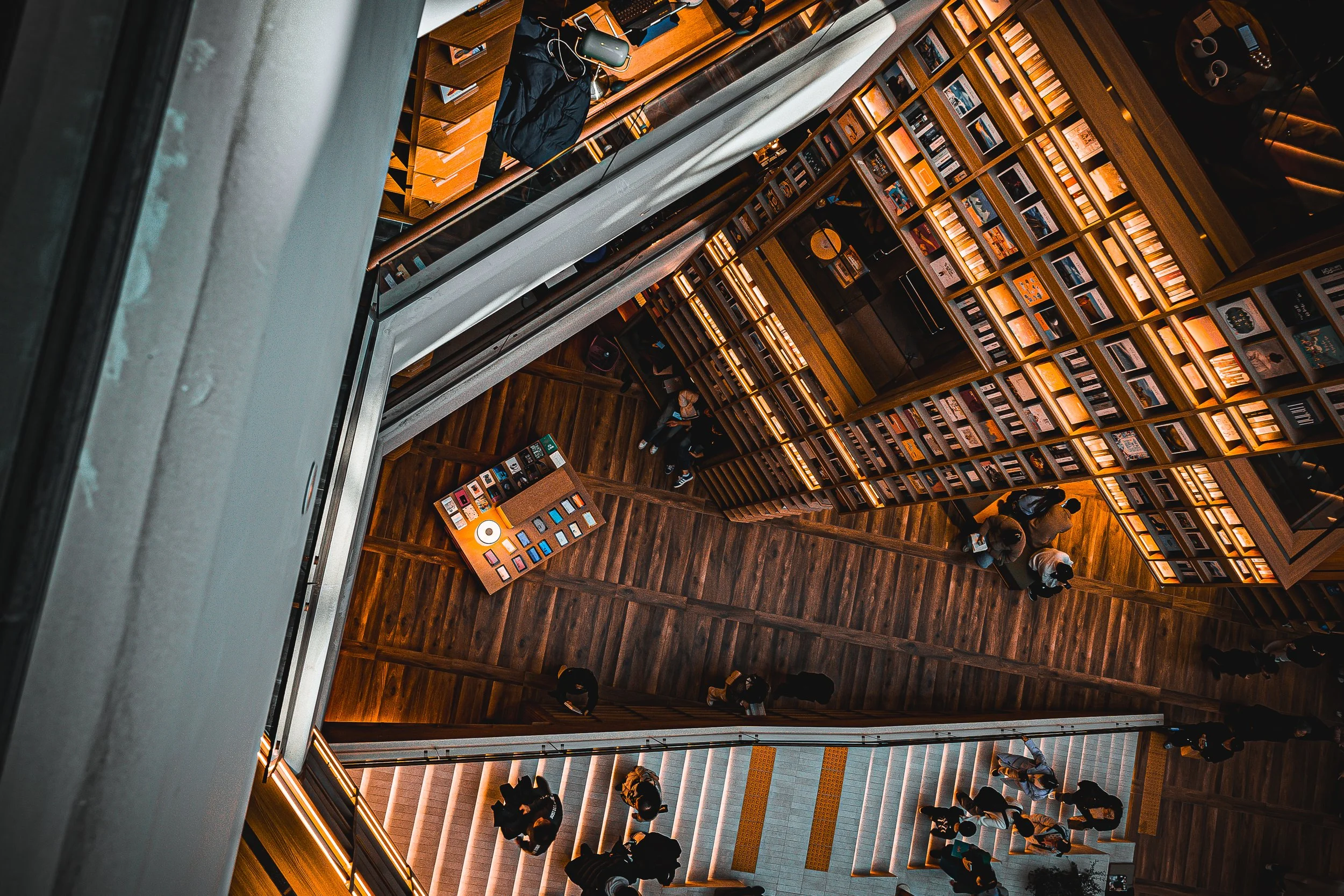 Interior view of a bookstore or library from above, showing shelves filled with books, a wooden floor, and a small table with books displayed. Several people are browsing and walking around.