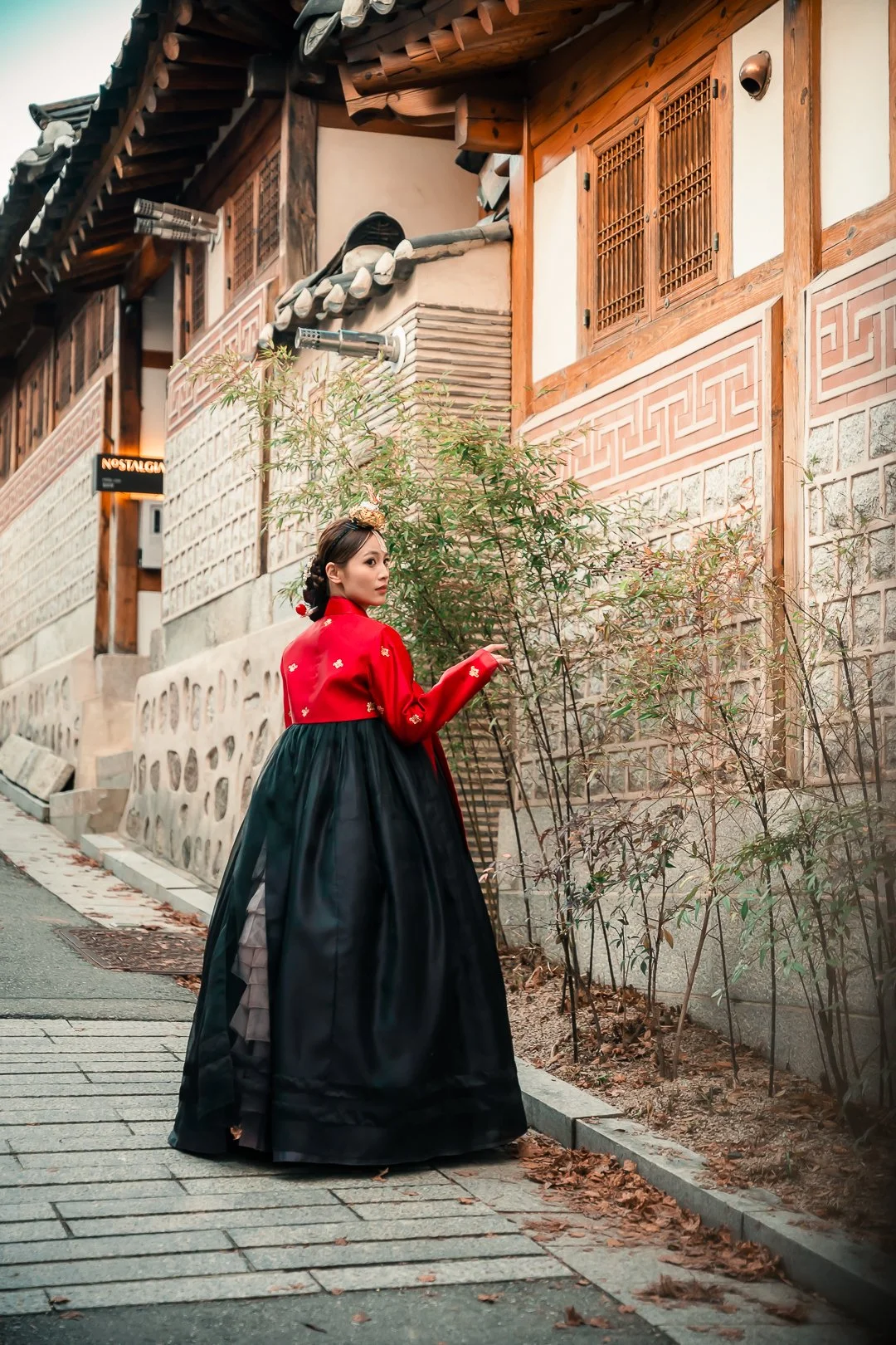 A woman dressed in traditional Korean hanbok attire standing on a street next to a bamboo plant, with traditional Korean house structures in the background.