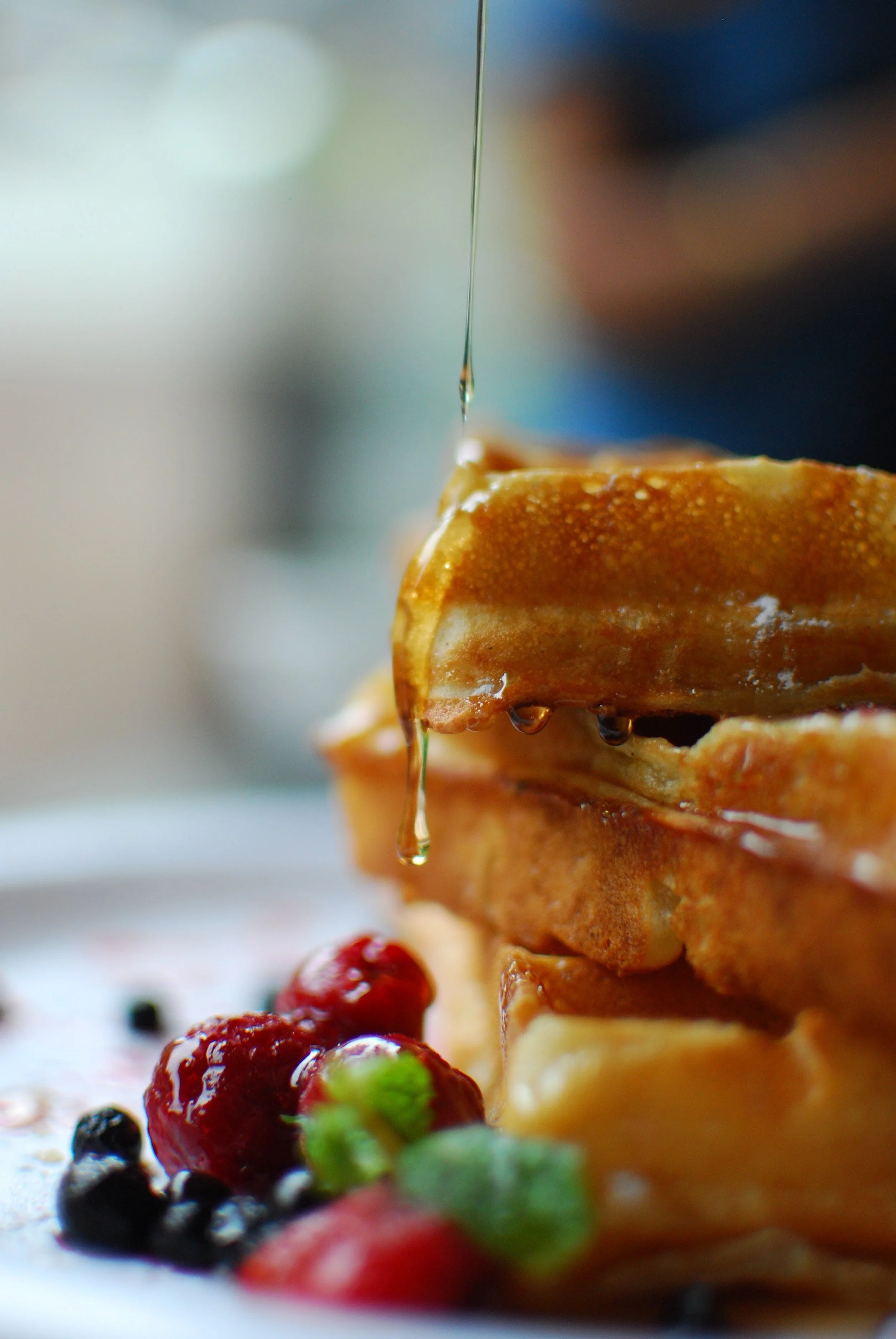 Close-up of stacked waffles with syrup being poured on top, surrounded by fresh berries.