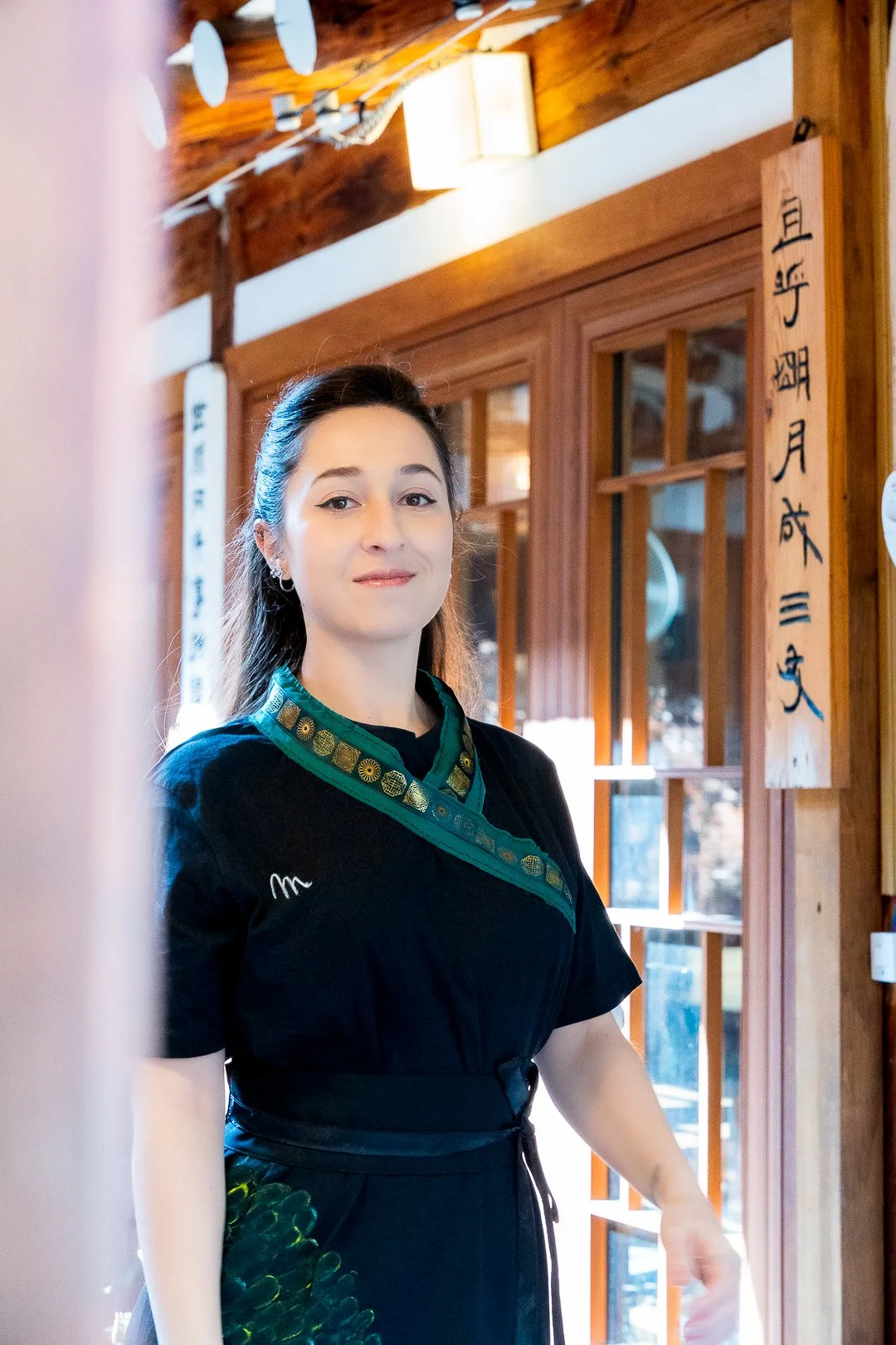 A woman with dark hair, wearing a black dress with green patterned accents, standing inside a wooden building with Japanese calligraphy on the walls.