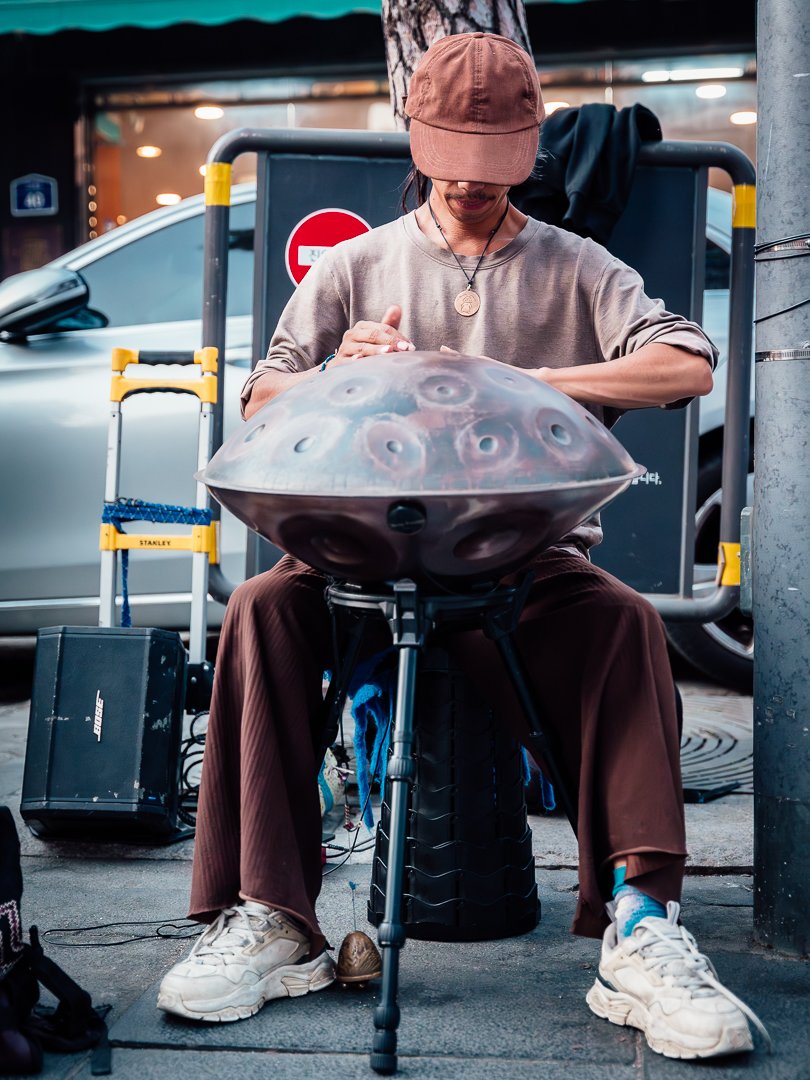 A person wearing a tan cap and brown pants is sitting outdoors on a stool, playing a steel tongue drum with their head slightly bowed. They are dressed casually and are surrounded by some equipment, with a city street in the background.