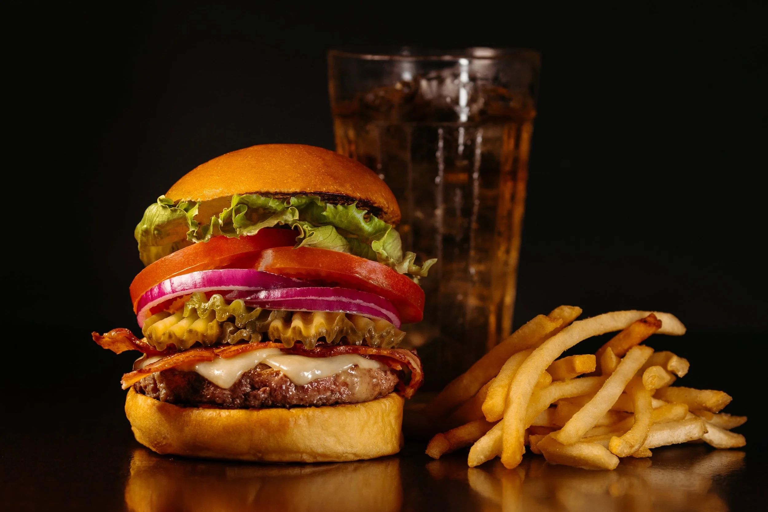 A cheeseburger with lettuce, tomato, pickles, onions, bacon, and cheese in a bun, served with a side of French fries and a glass of soda on a dark background.