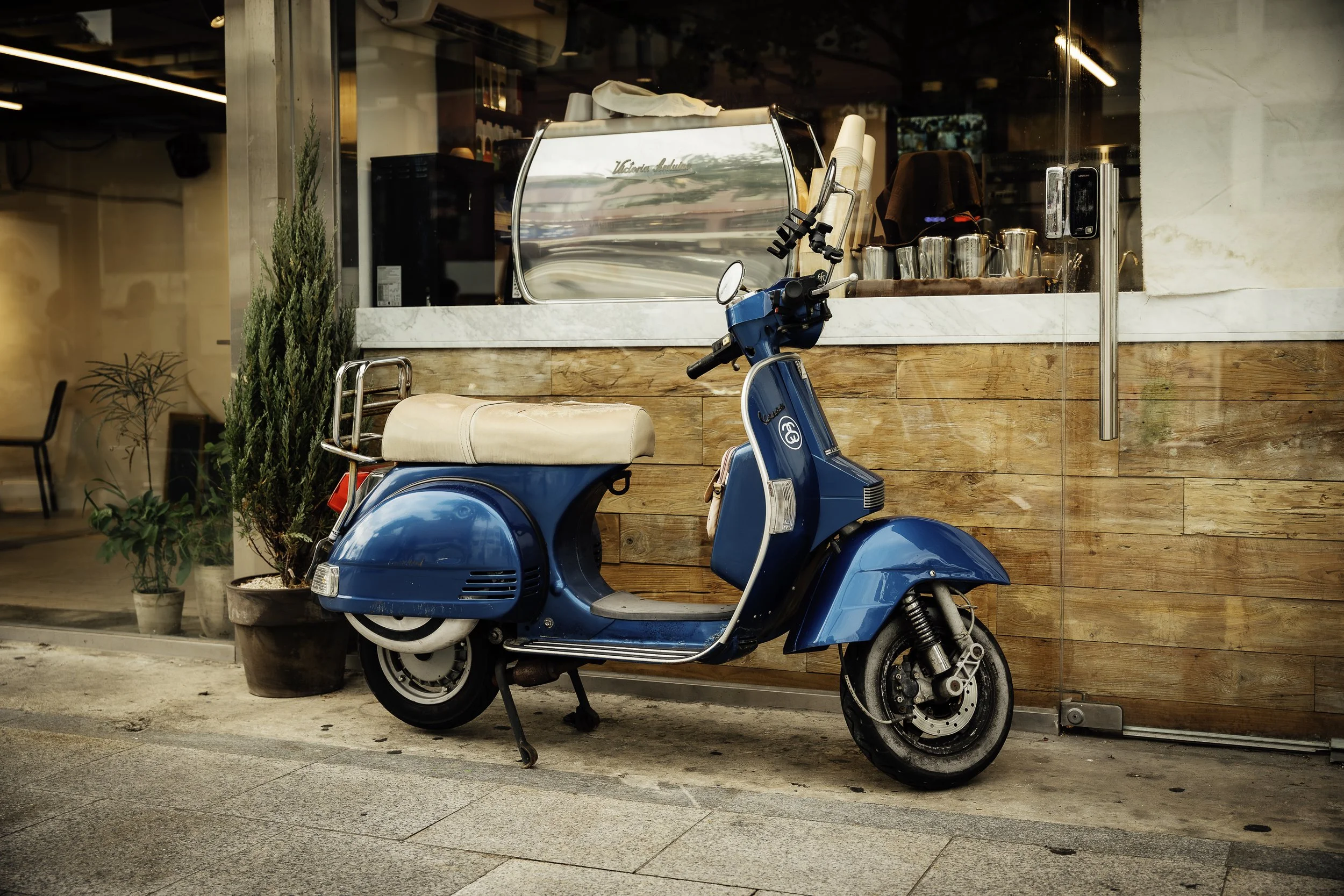 Blue vintage scooter parked outside a coffee shop with wooden wall, potted plants, and a large window with cafe equipment visible inside.