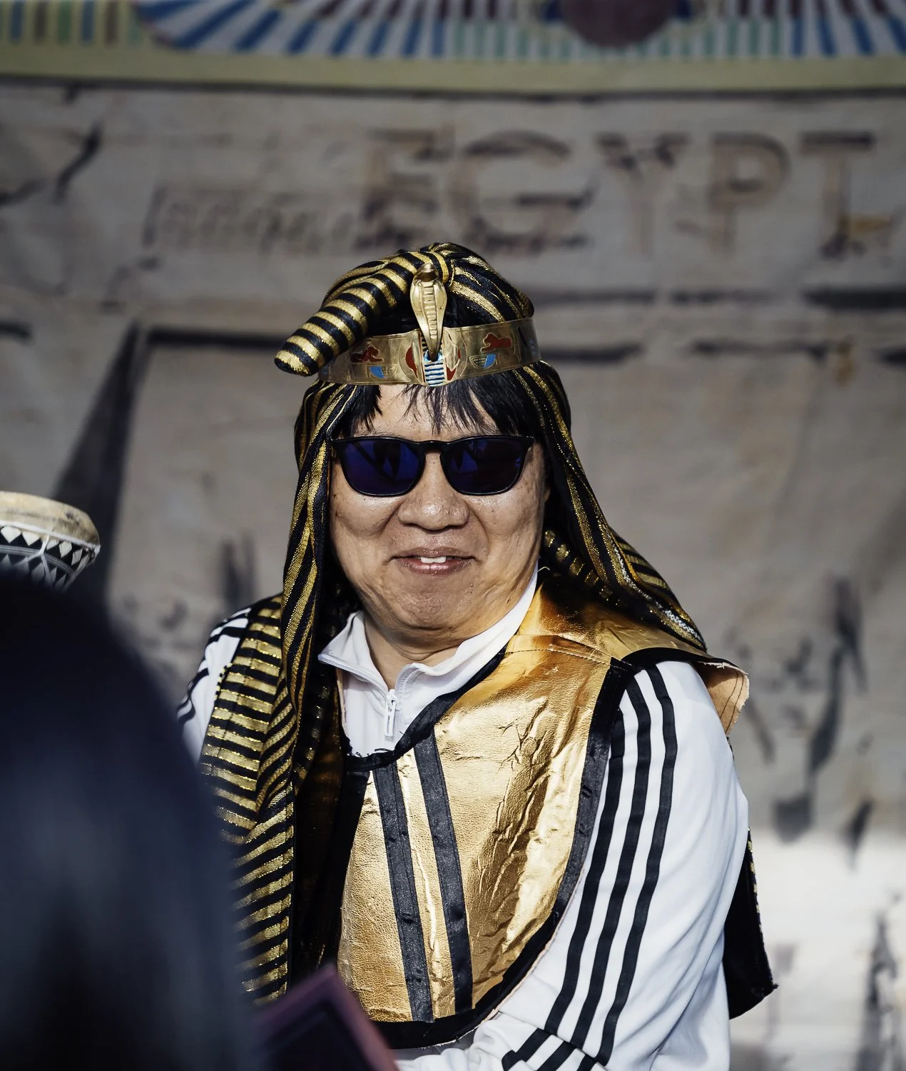 A person dressed in Egyptian-themed costume, wearing a headdress with a cobra, a golden vest, sunglasses, and a white sports jacket with black stripes, standing in front of a backdrop with some text and symbols.