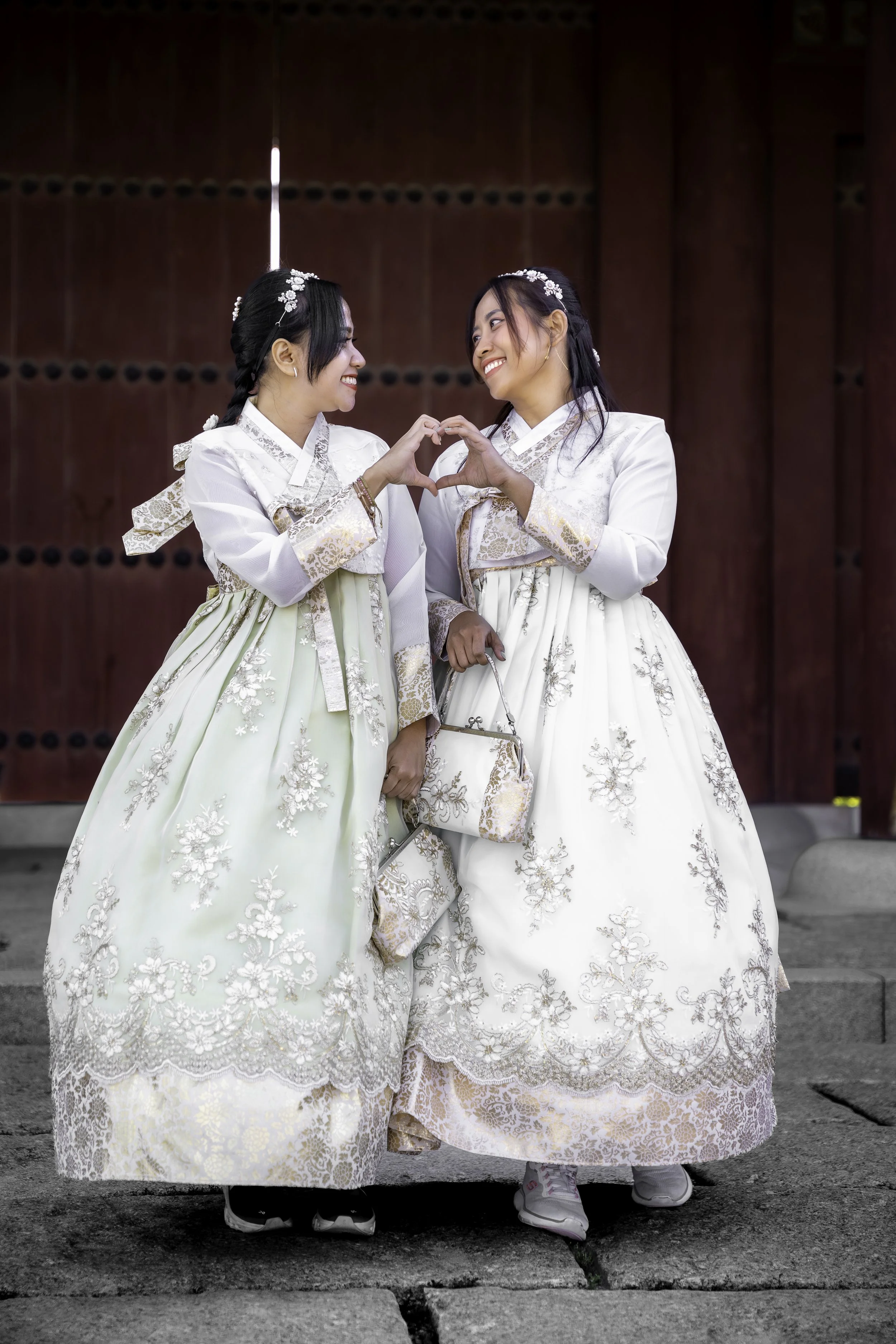 Two women wearing traditional Korean dresses, hanbok, standing in front of a large wooden gate, smiling and forming a heart shape with their hands.