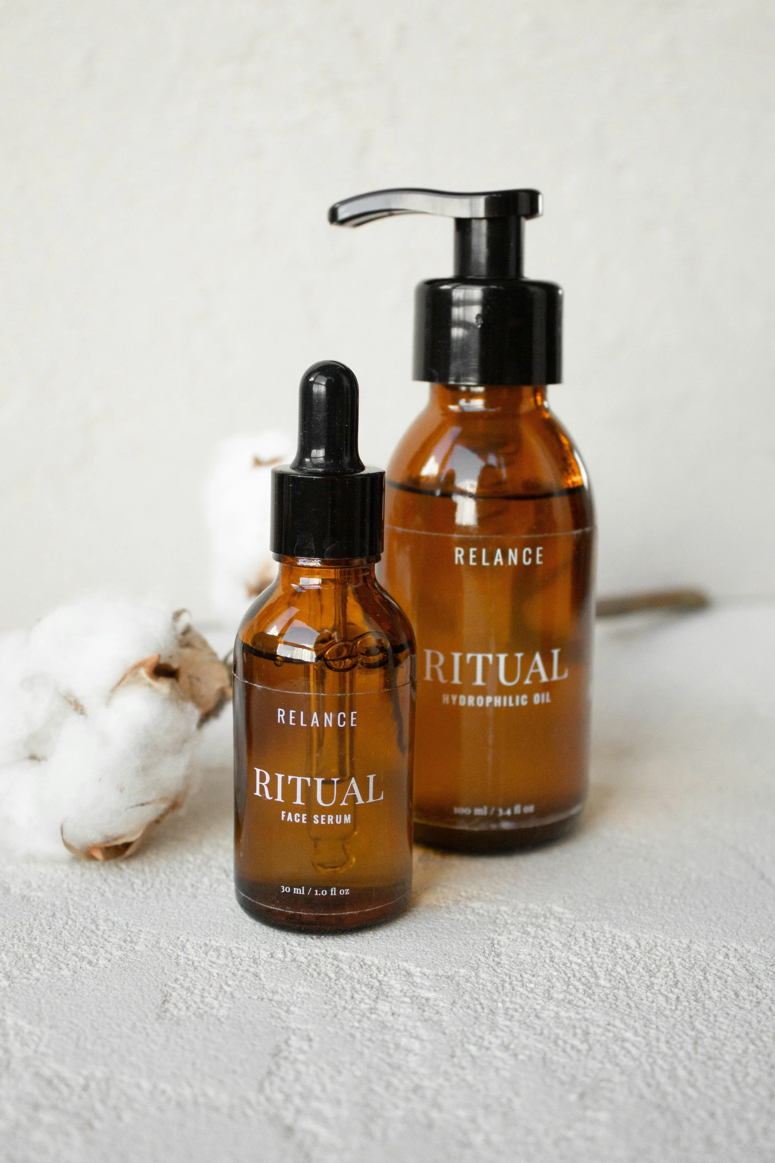 Two amber glass bottles with black caps and pumps labeled 'Ritual,' one for face serum and the other for hydrophilic oil, placed on a textured white surface with a piece of cotton next to them.