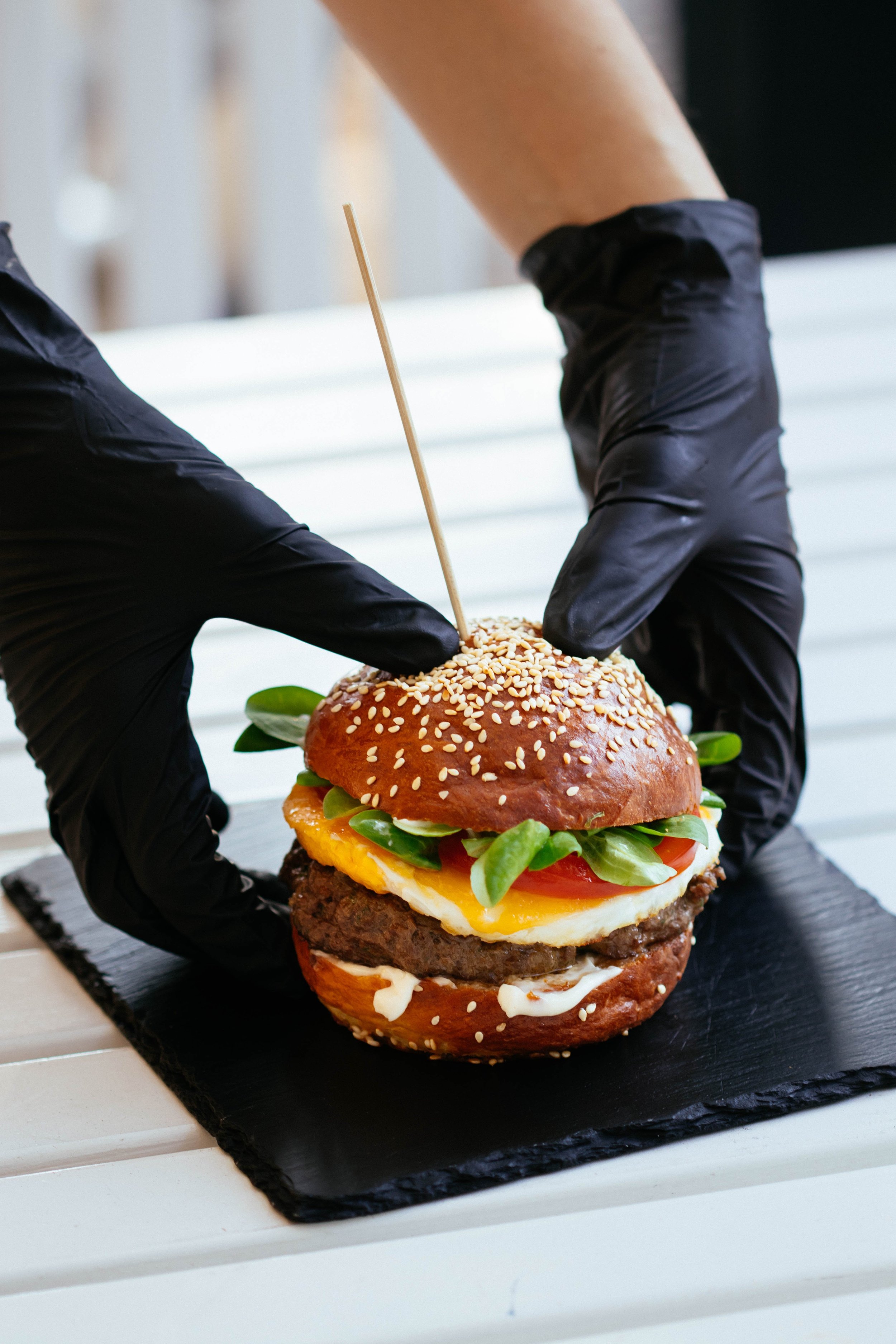 Person wearing black gloves assembling a large gourmet burger with sesame seed bun, lettuce, tomato, fried egg, beef patty, and sauces.