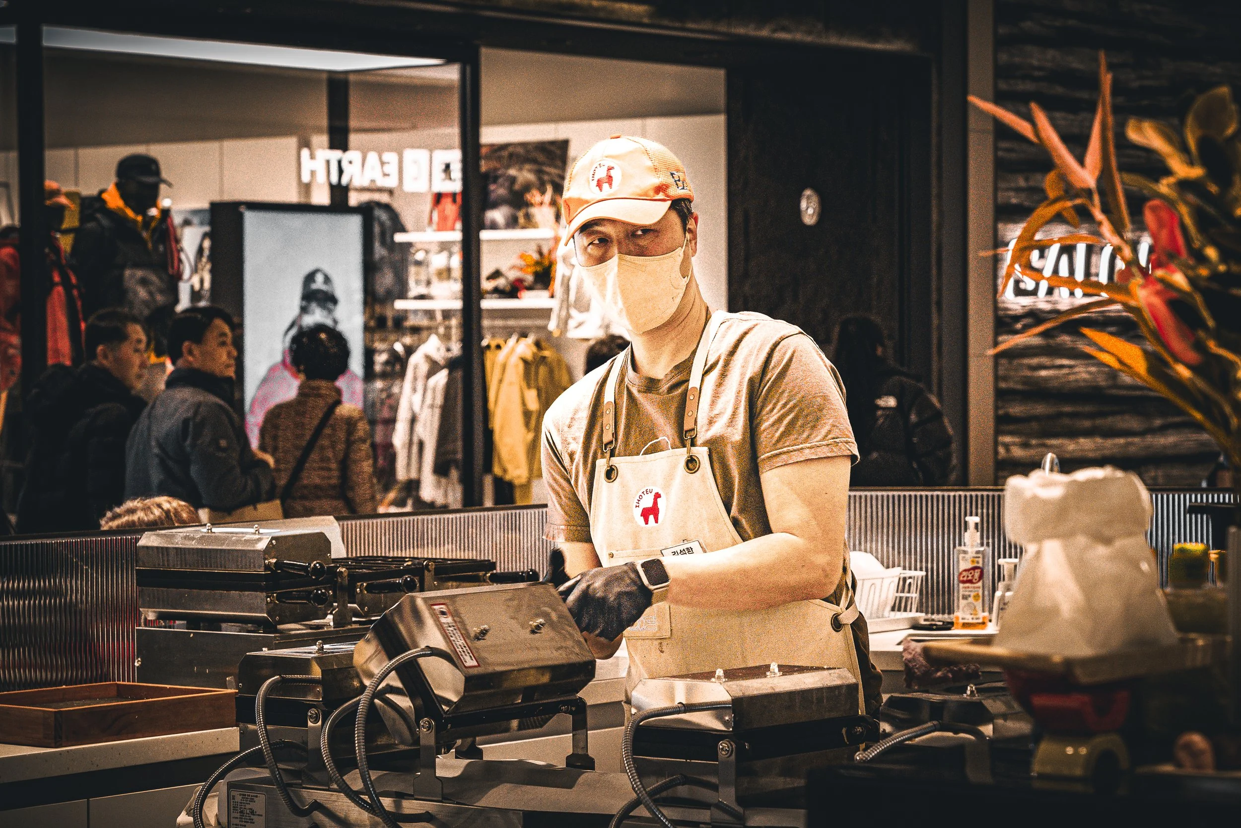 A man wearing a beige apron, face mask, and cap working at a food stand in a busy indoor location, with customers and clothing racks visible in the background.