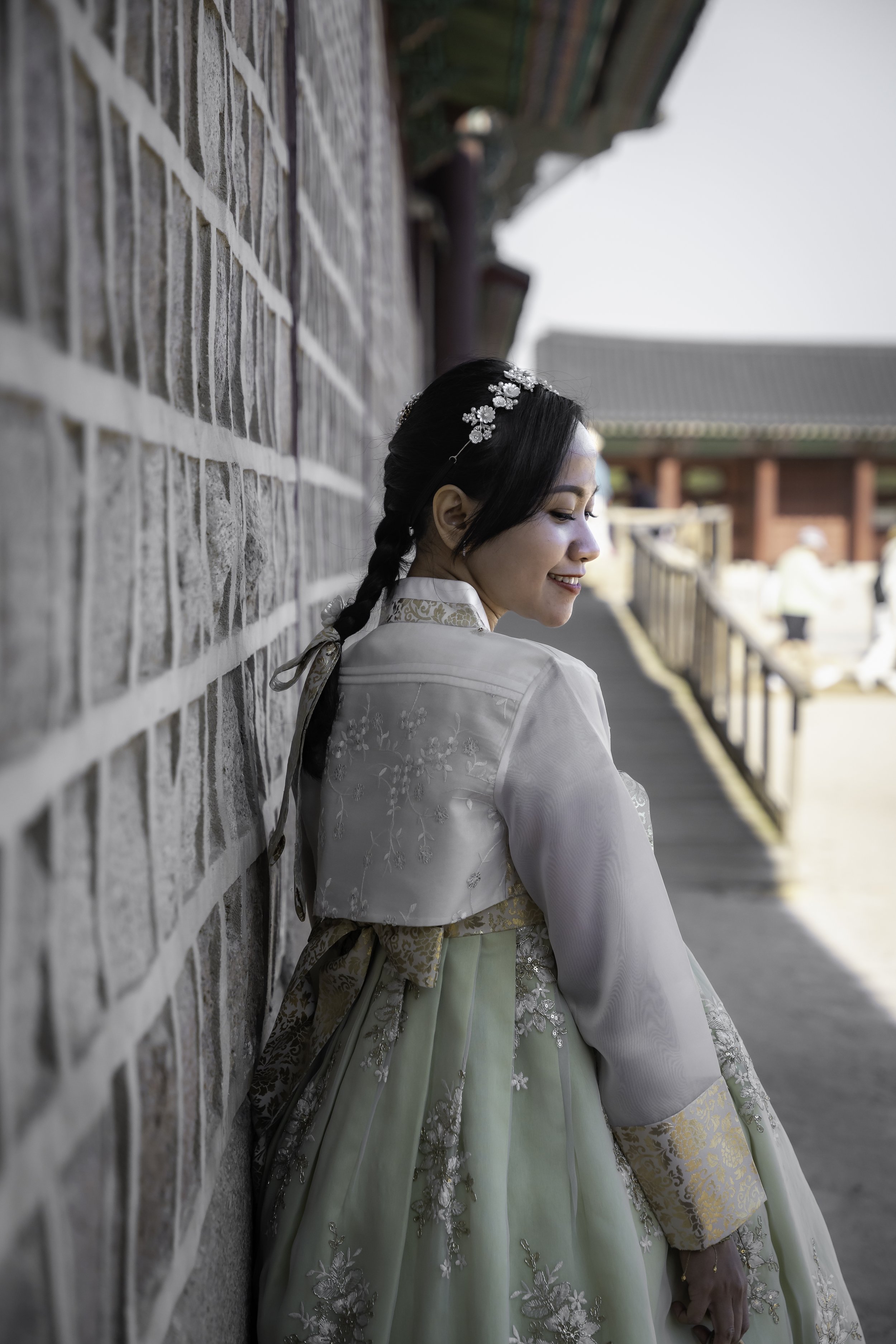 A young woman with dark hair styled in a braid, wearing a traditional Korean hanbok, smiling and leaning against a stone wall with traditional architecture in the background.