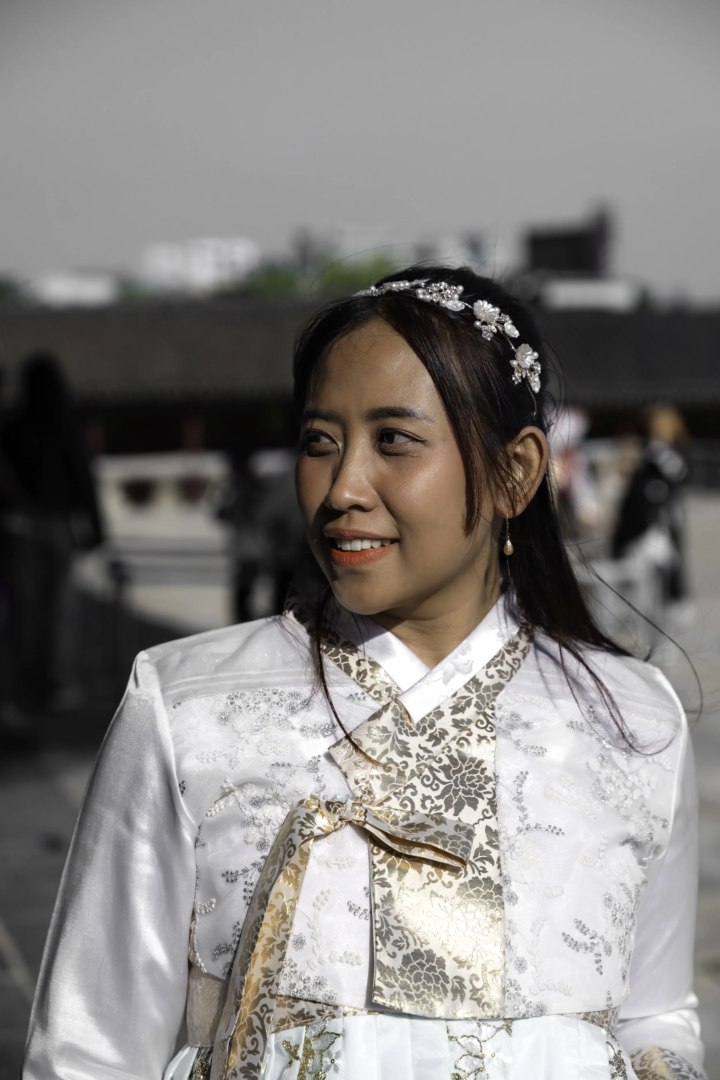 A woman in traditional Korean hanbok attire smiling and looking to her left outdoors on a sunny day.