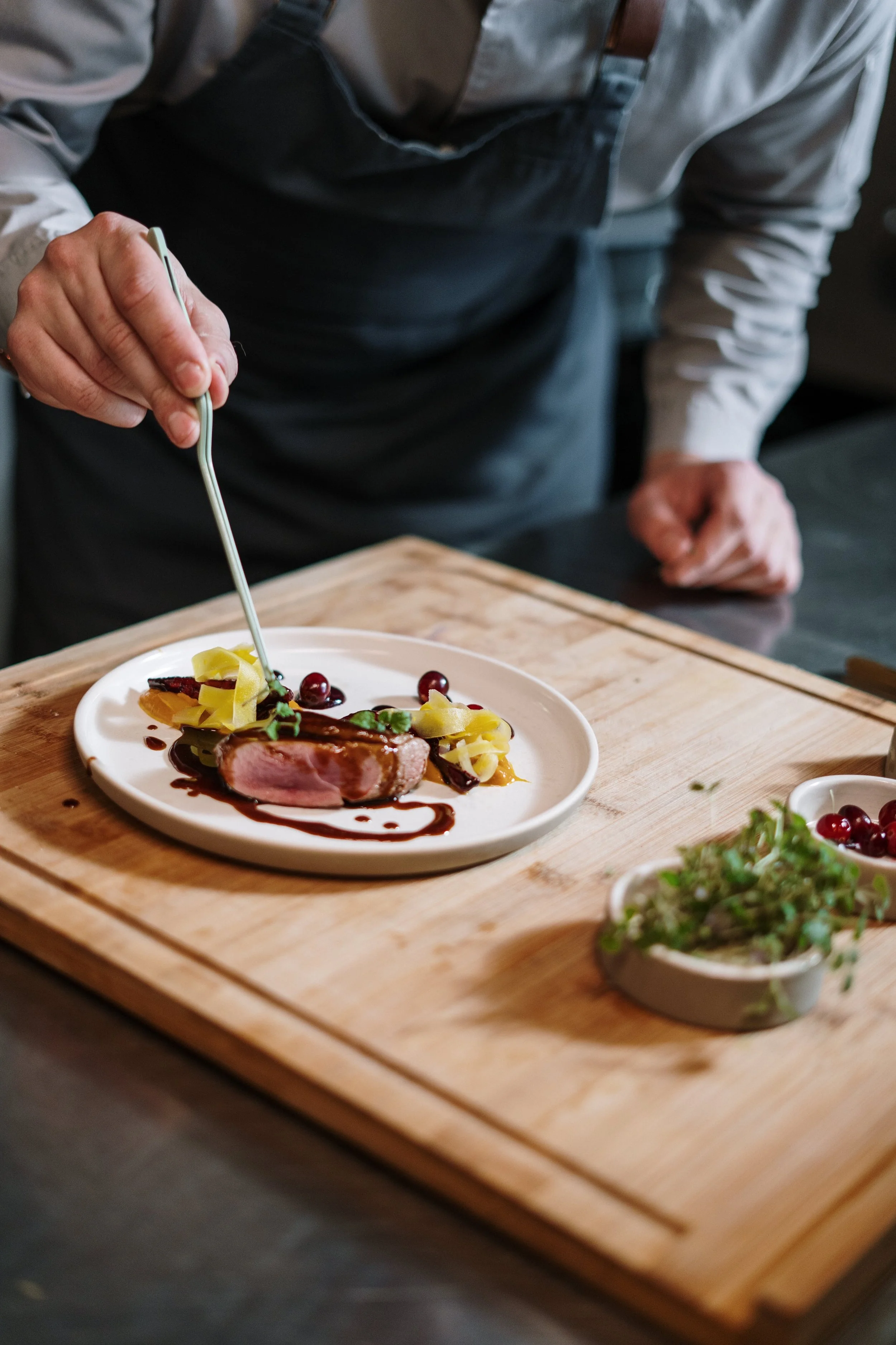 Chef garnishing a plated dish of seared meat with sauce, accompanied by vegetables and garnishes on a white plate.