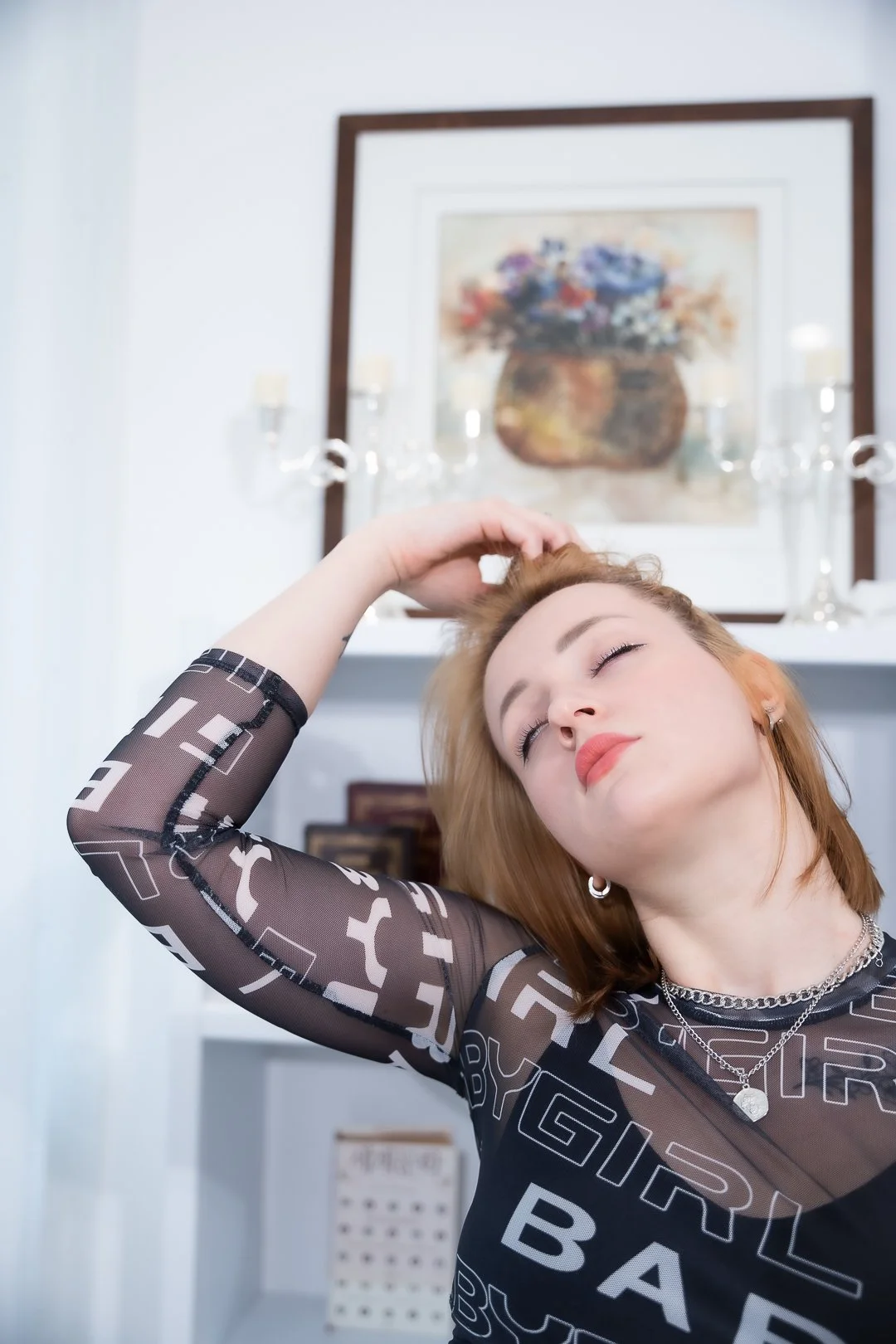 Young woman with red hair and closed eyes, wearing a black sheer top with white text, posing with her right hand on her head, in a room with a framed flower painting and candles in the background.