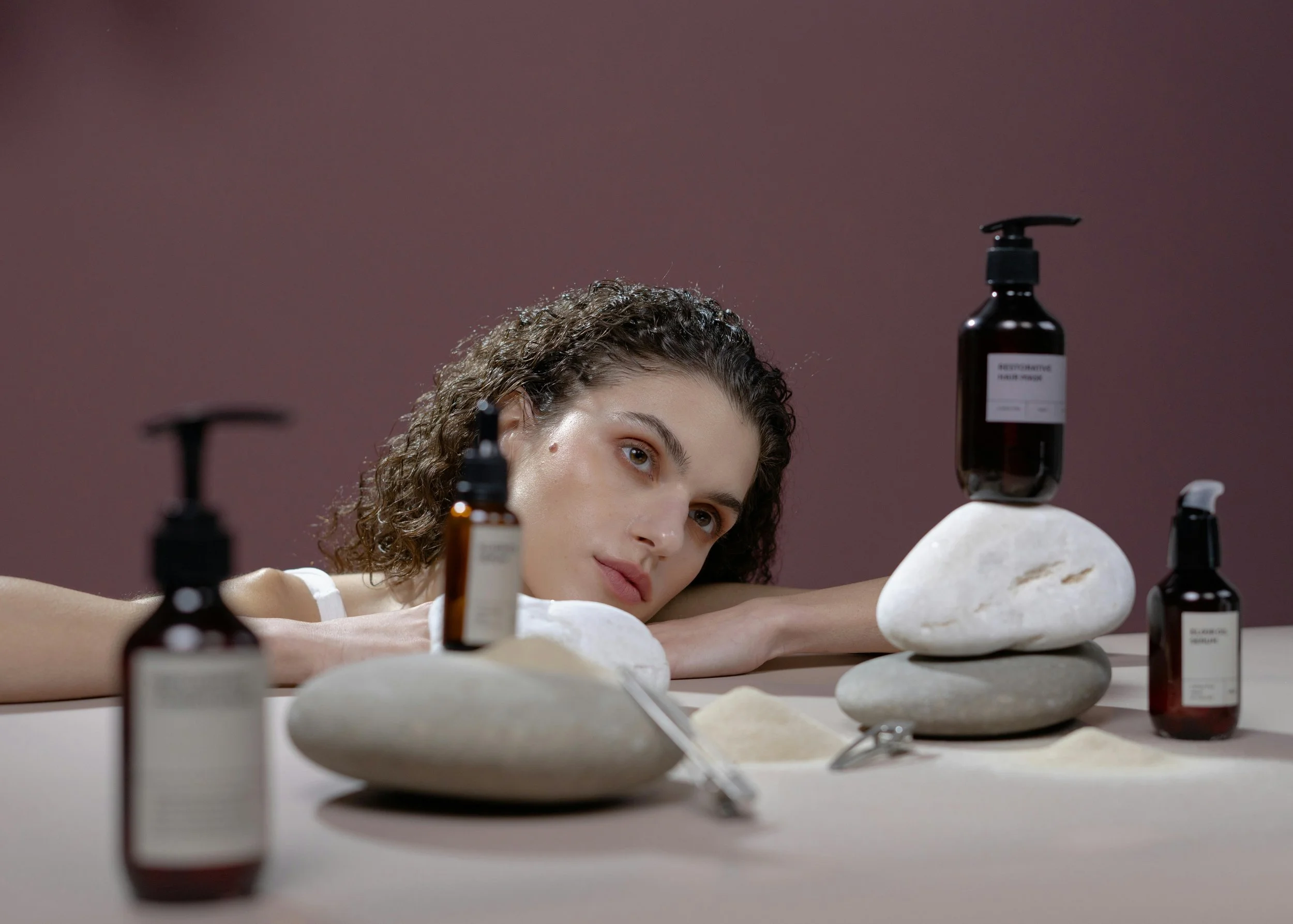 A woman with curly hair laying on her side with her chin resting on her arm, surrounded by bottles and stones, in a calming, minimal setting with a mauve background.