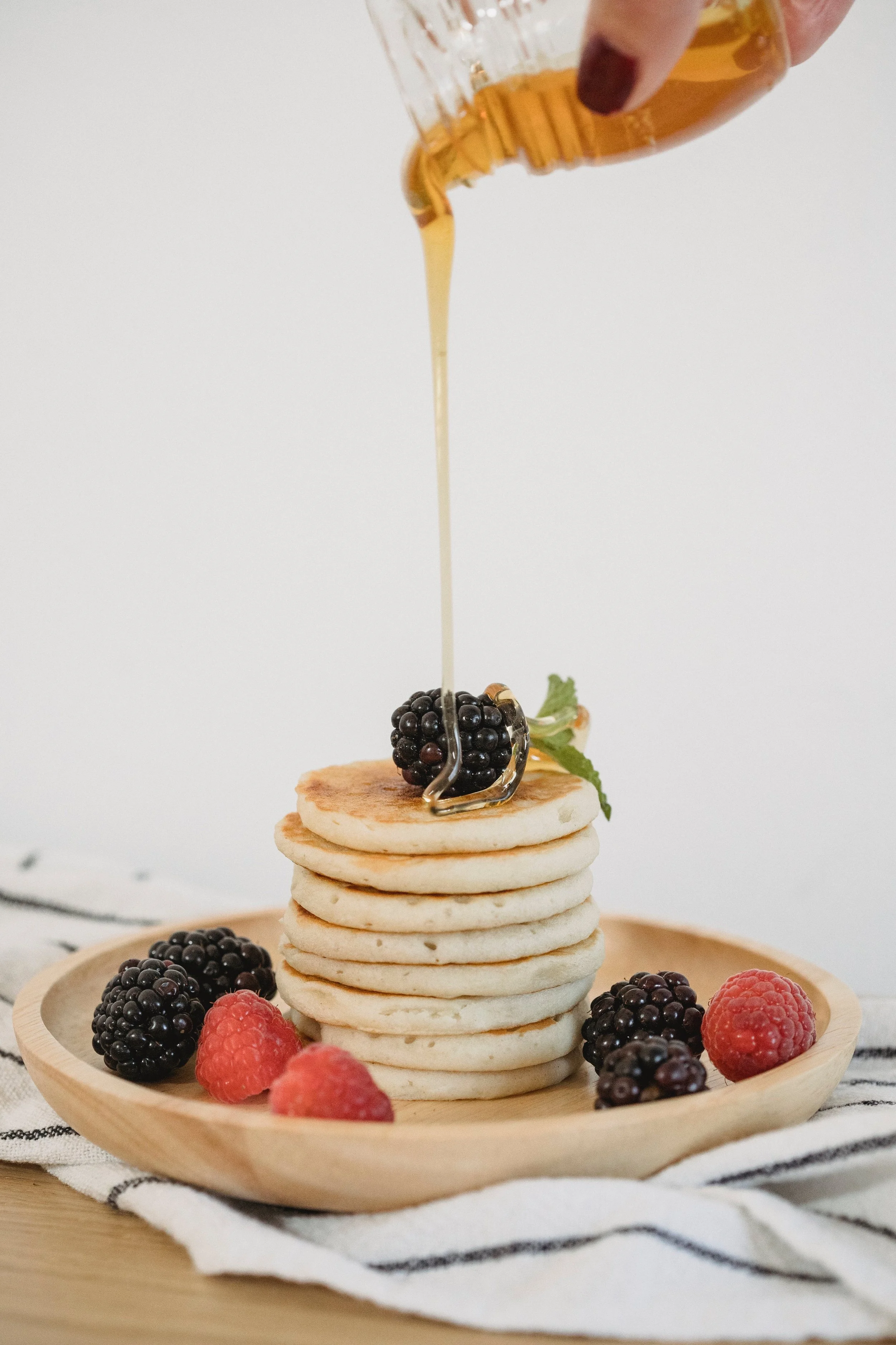 Stack of six pancakes topped with blackberries and a sprig of mint on a wooden plate, with more berries around. Syrup is being poured over the pancakes from a glass container.