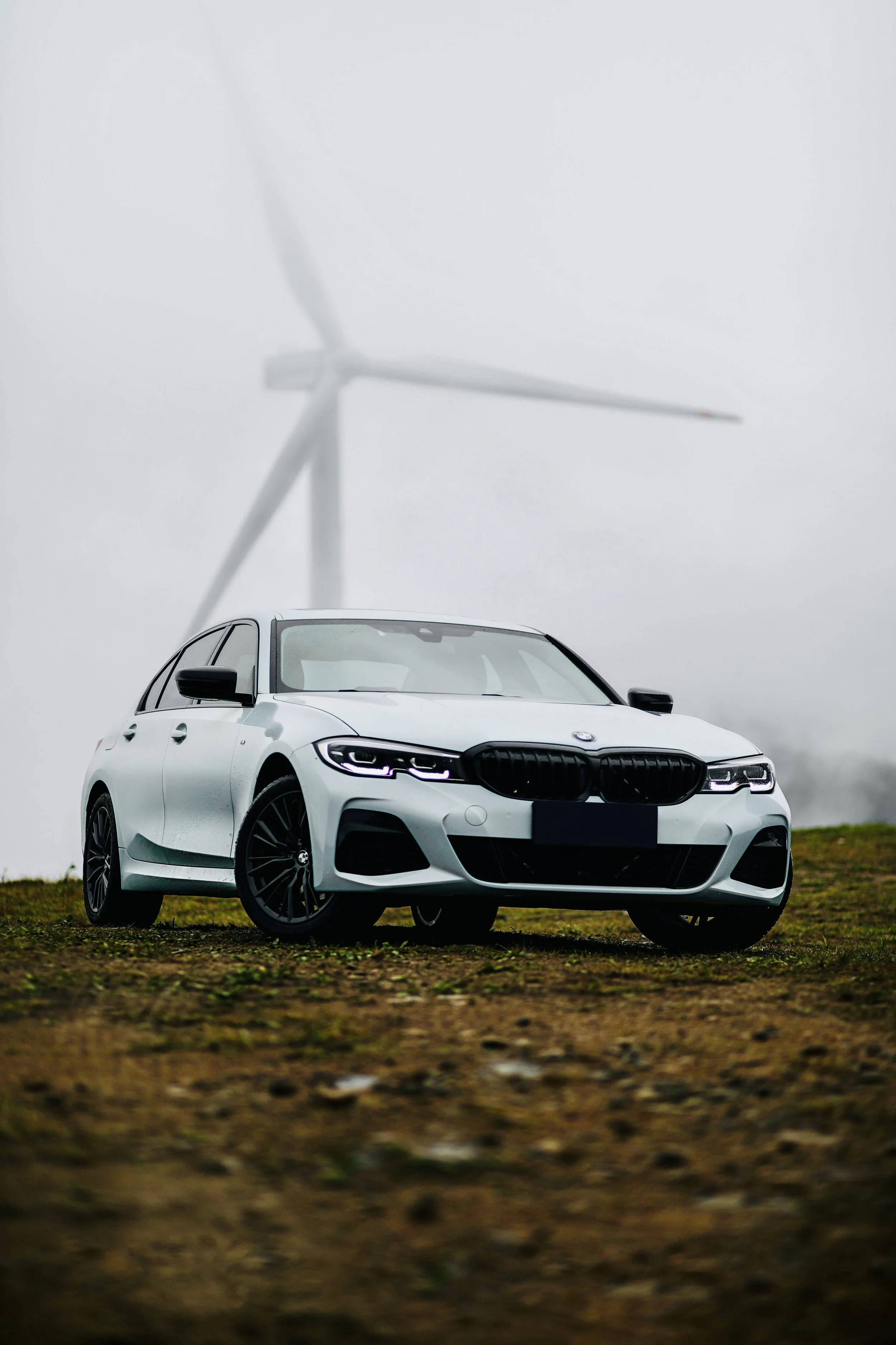 White BMW car parked on grassy terrain with a wind turbine in the background on a cloudy day.
