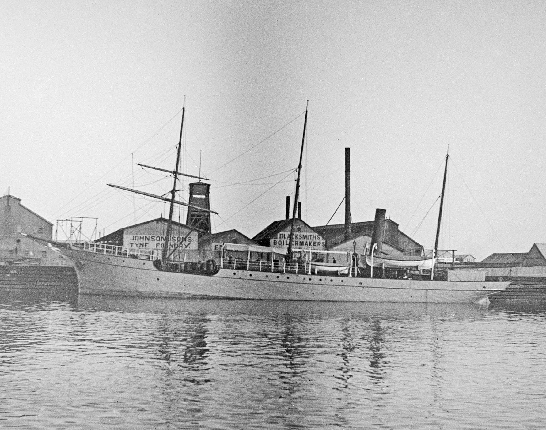 Black and white photo of a boat docked in a harbor with buildings in the background, some with signs reading "Johnson's Spas" and "Blacksmiths, Boiler Makers."