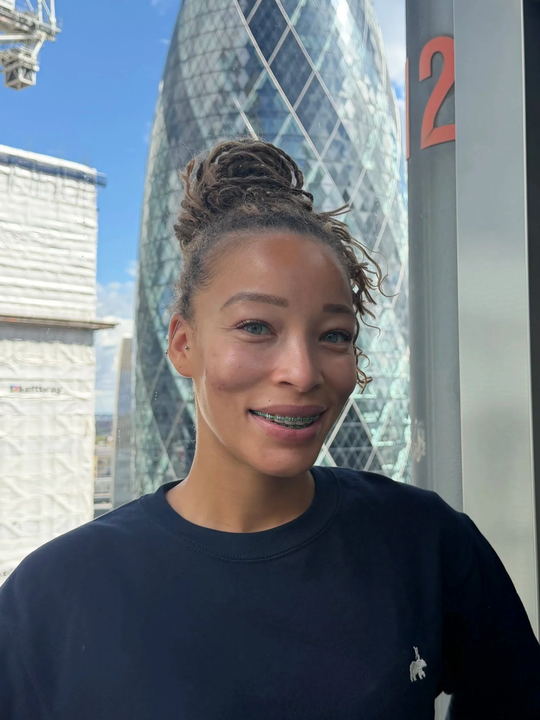 Close-up of a young woman with shoulder-length dark hair, smiling softly, wearing a navy blue sweatshirt with a small white logo of a rhinoceros on the left side, against a blurred cityscape background with tall buildings.