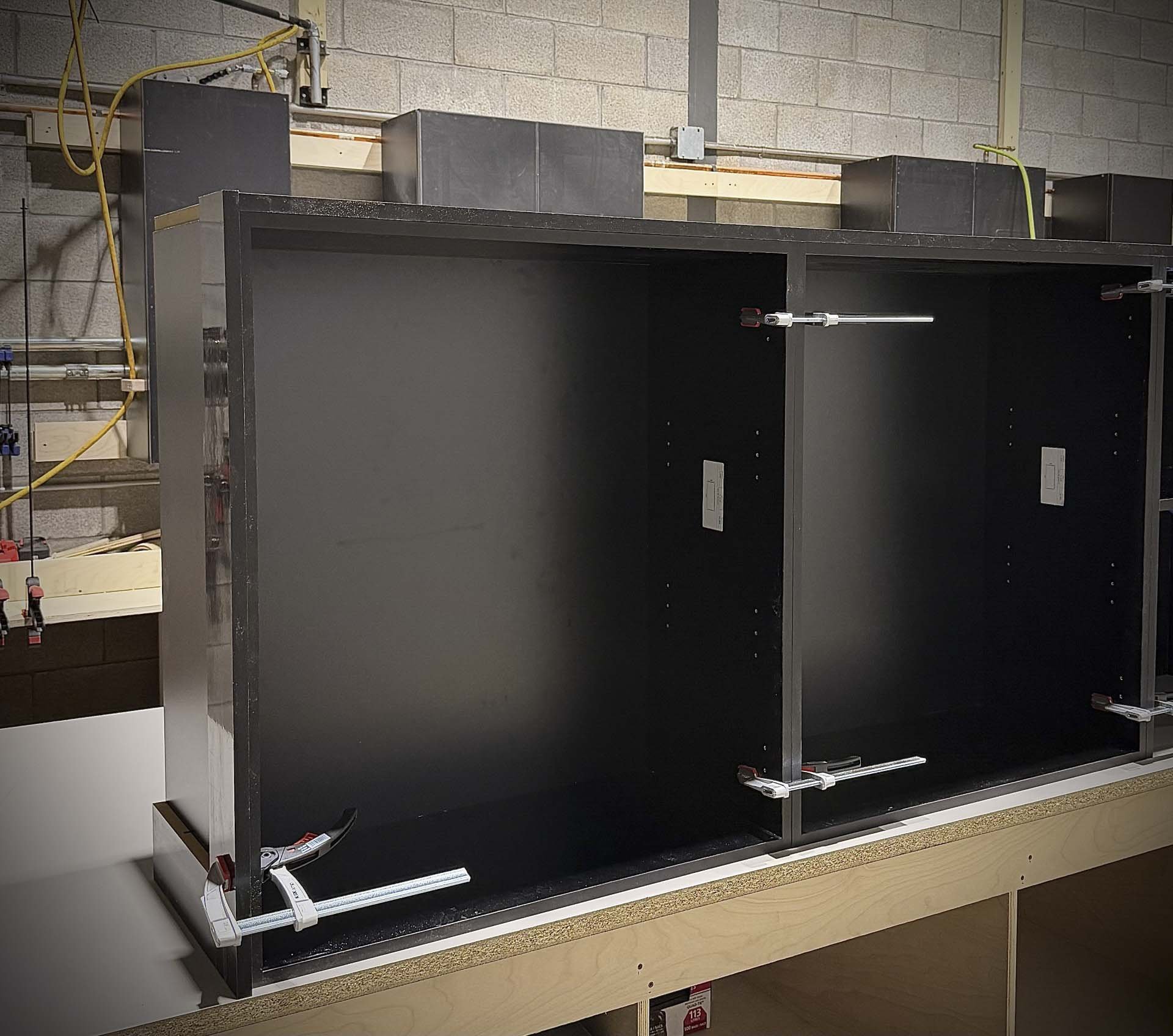 Fabrication en atelier de caissons d'armoires noirs sur mesure assemblés avec des serres-joints pour projet d'ébénisterie architecturale et commerciale à Boisbriand.