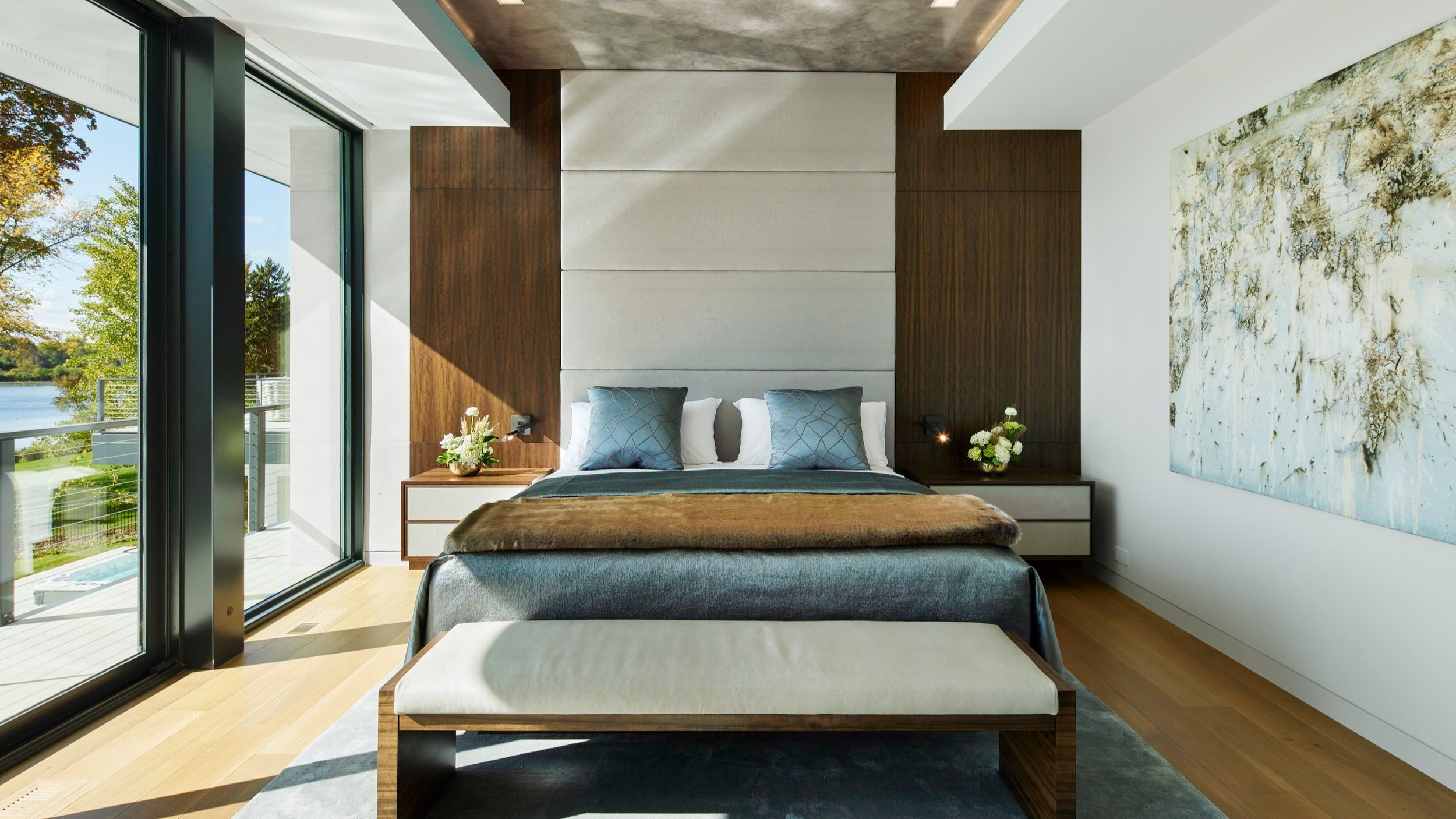A warm modern bedroom with floor-to-ceiling windows, recessed lighting, and beautiful artwork