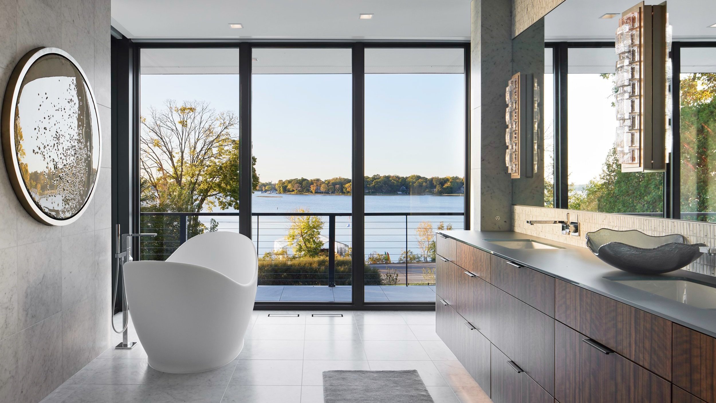 A warm modern bathroom that features floor-to-ceiling windows with a view of the lake, a freestanding bathtub, and refined modern finishes