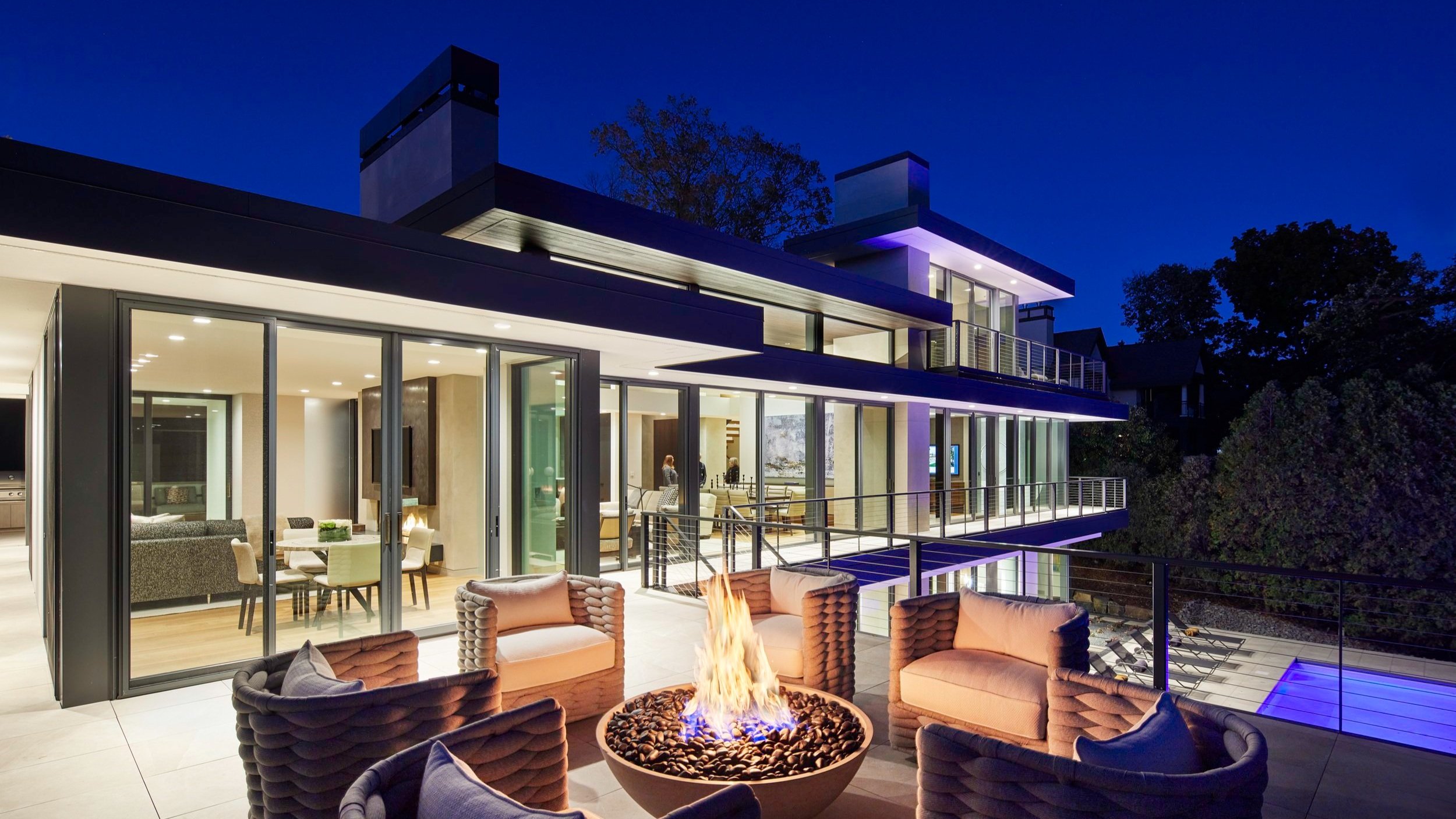 A warm modern lake home with floor-to-ceiling windows at dusk. A glowing fire is lit on the patio of this second-level balcony, which contrasts beautifully with the deep blue sky above.