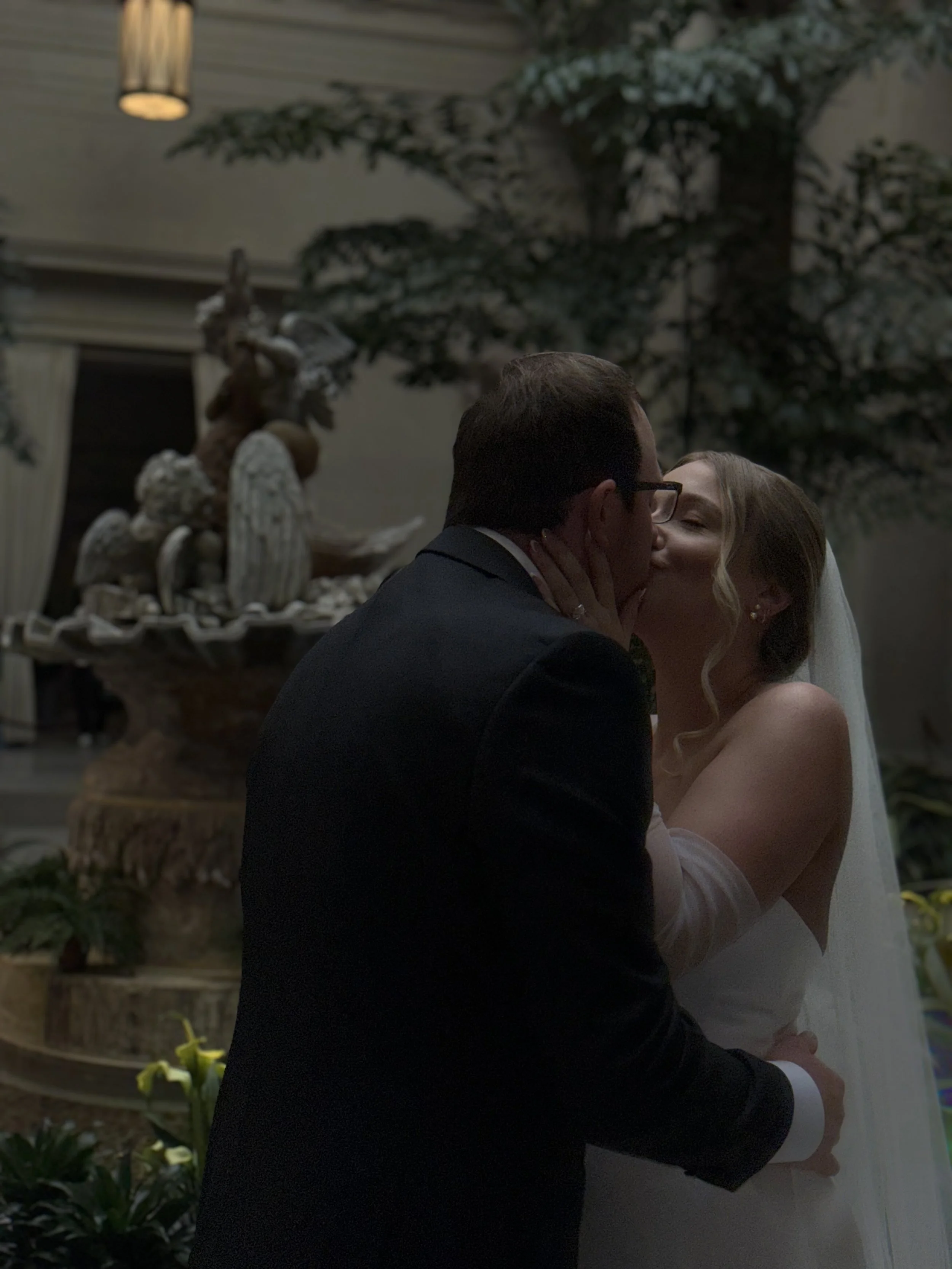 A bride and groom sharing a kiss at their wedding, with a decorative fountain and greenery in the background.