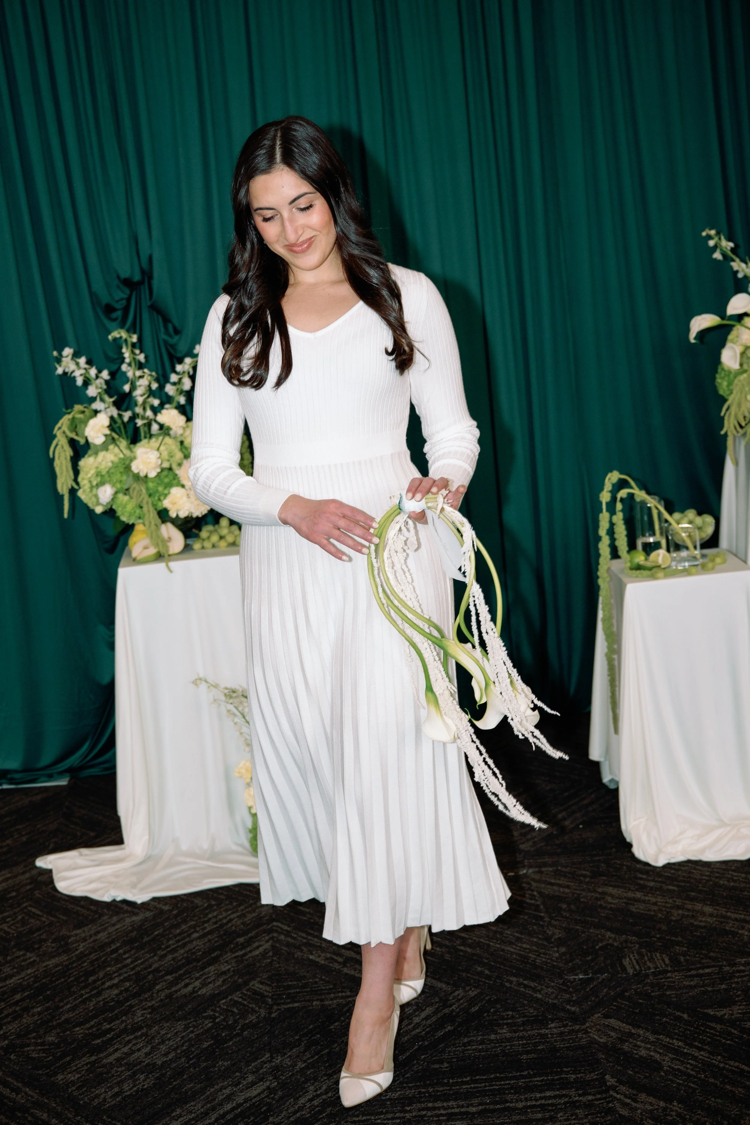 A woman in a white pleated dress holding a white calla lily flower arrangement at a formal event with green curtains and floral decorations in the background.