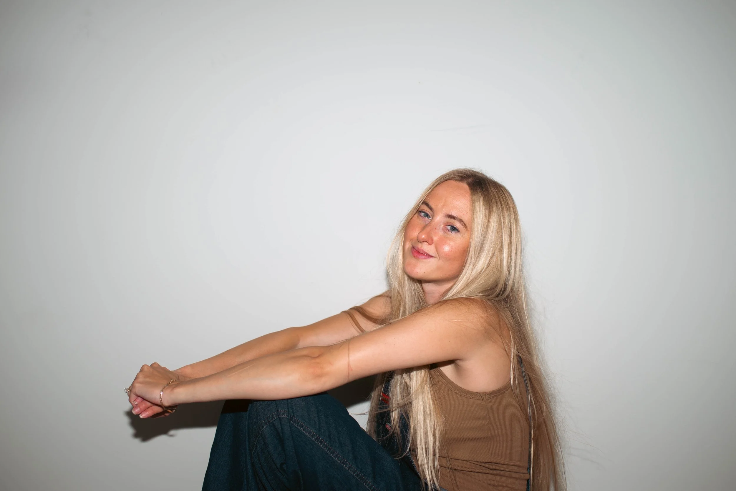 A young woman with long hair smiling outdoors in a grassy field, wearing a black top and light-colored jeans, with bare trees in the background.