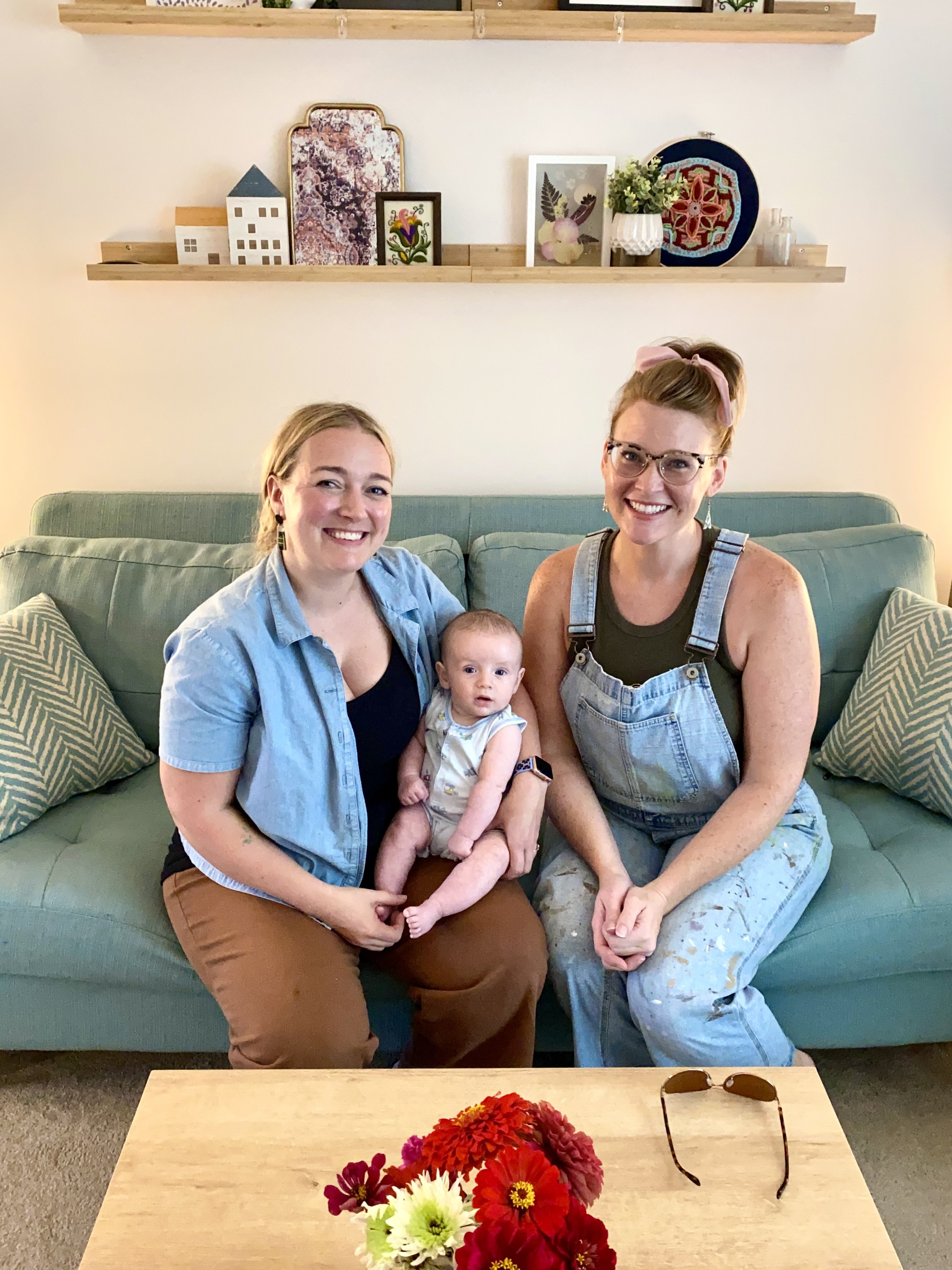 A photo of Mary Foltz of Ambrosia Lifecasting sitting on a teal sofa in her home studio with a Mother and child that are clients.