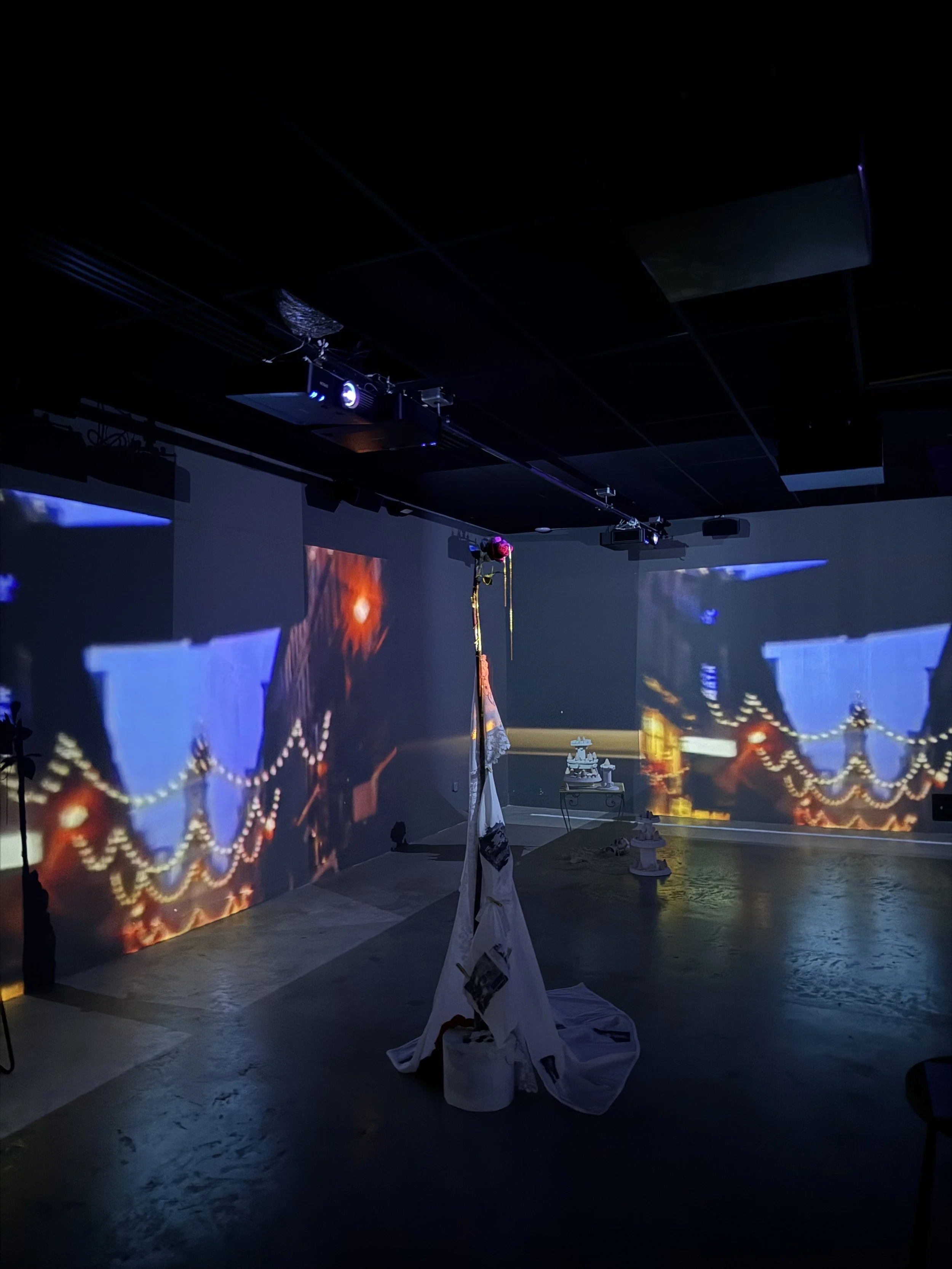 Dark room with projected images of a bridge and city skyline on the walls, stage set with a colorful tent and hanging decorations, and a table with objects.