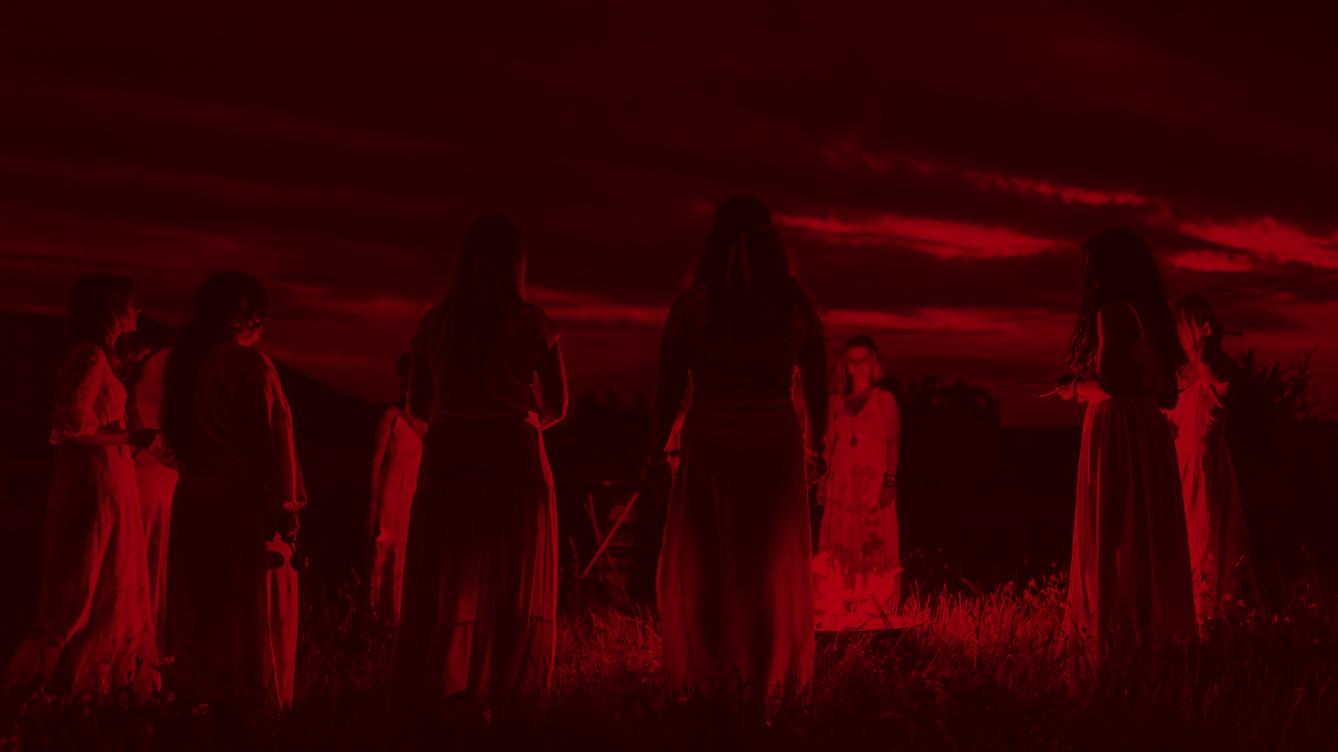 Group of people in long dresses gathered in a grassy field beneath a dramatic red sunset sky