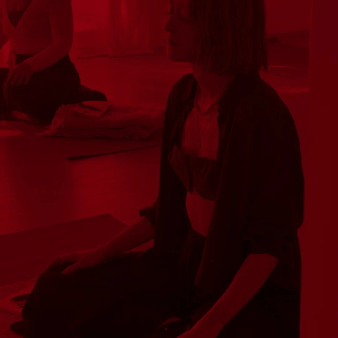 Person seated indoors in a dimly lit room, with another person and papers in the background