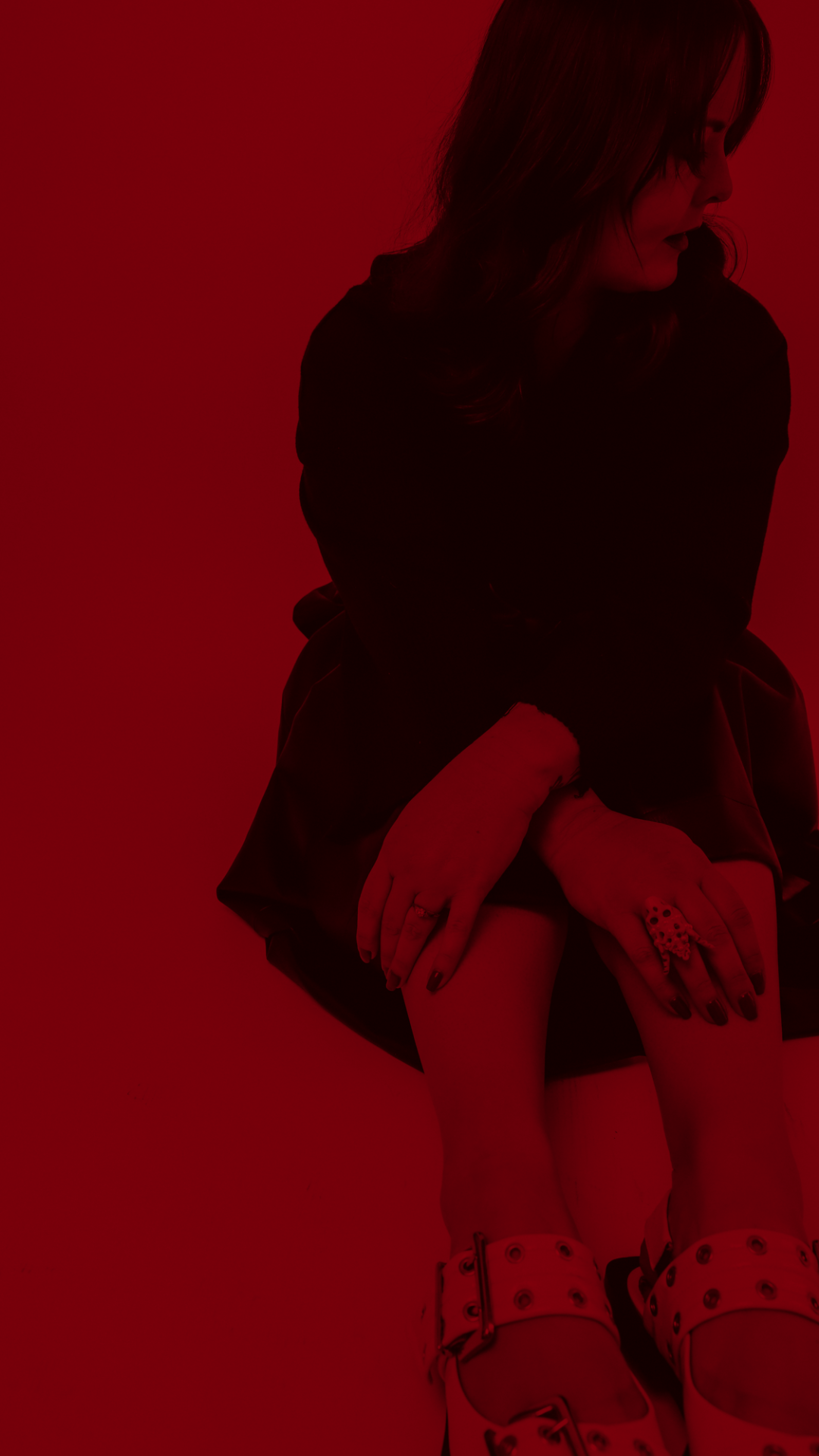 A woman with dark hair sitting on the floor, wearing a dark jacket and patterned high-heeled shoes, with her hands on her knees, against a red background.