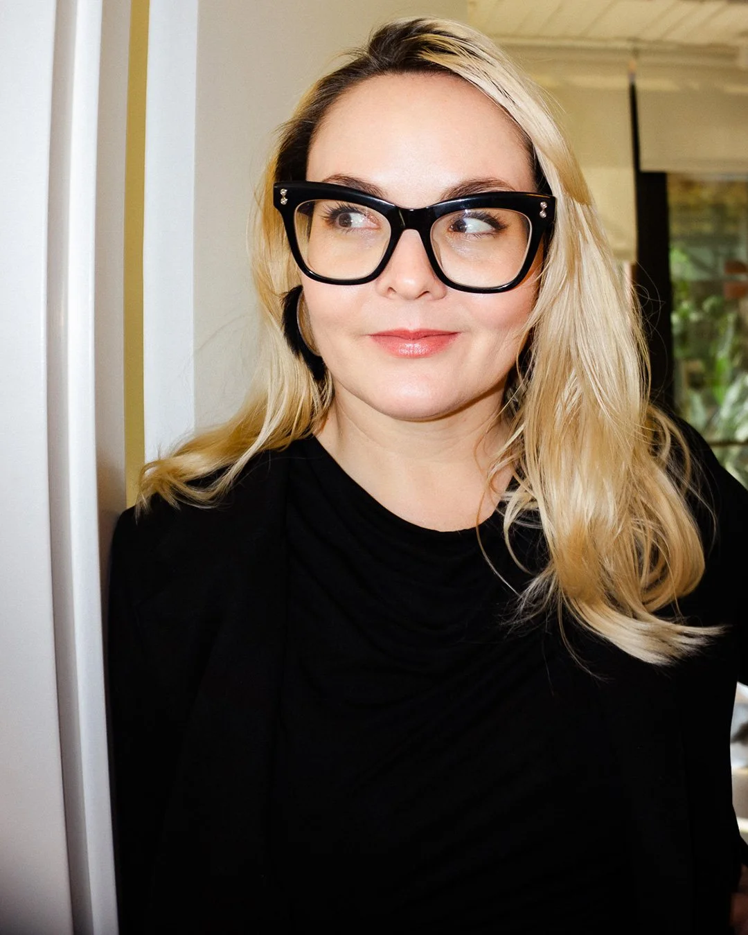 A woman with blonde hair wearing black glasses and a black top, looking to her left with a slight smile.