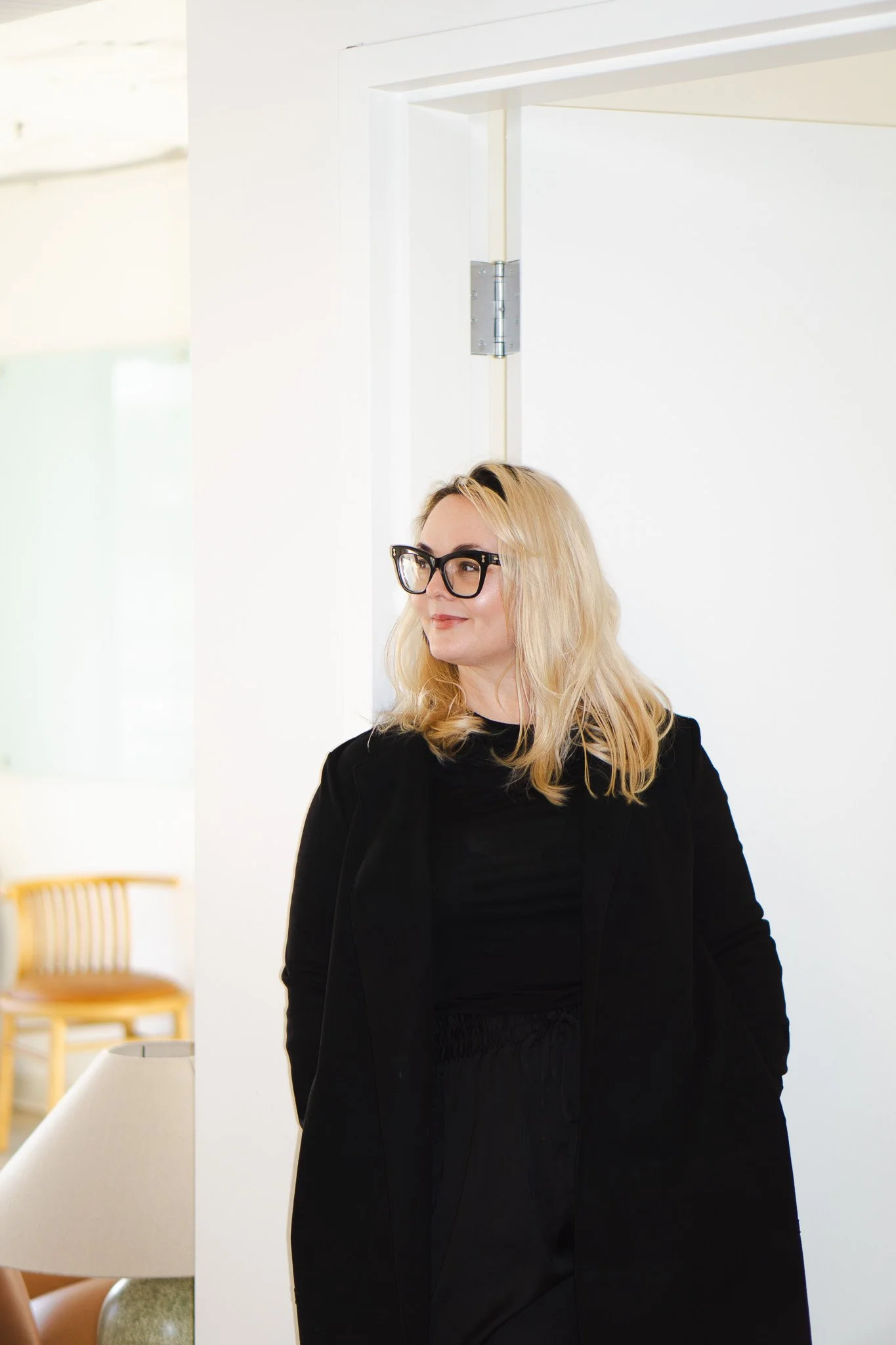 Woman with blonde hair and glasses wearing a black blazer standing indoors, white wall background, chair and lamp in the background.