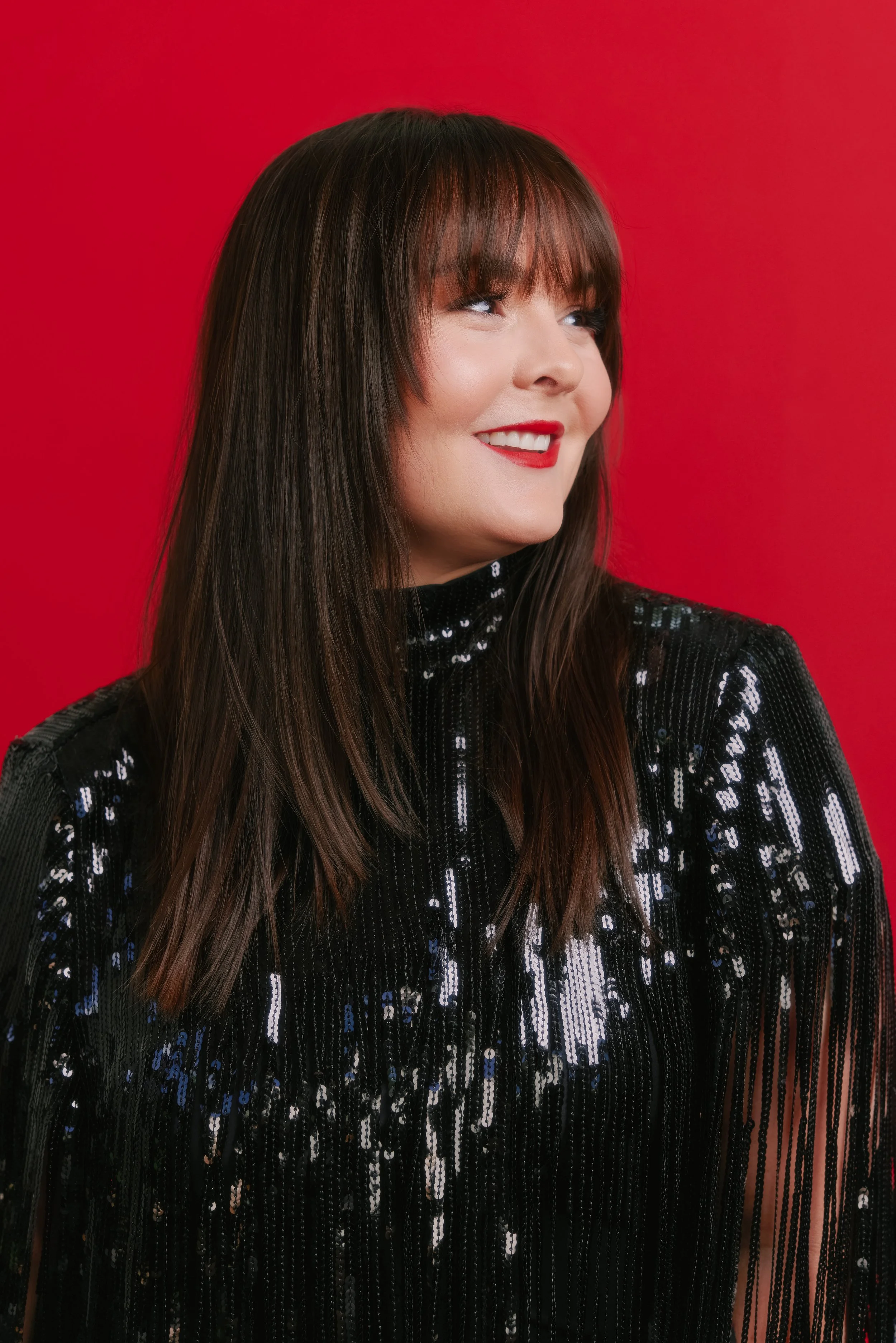 A smiling woman with long dark hair, red lipstick, wearing a black sequin top, against a red background.