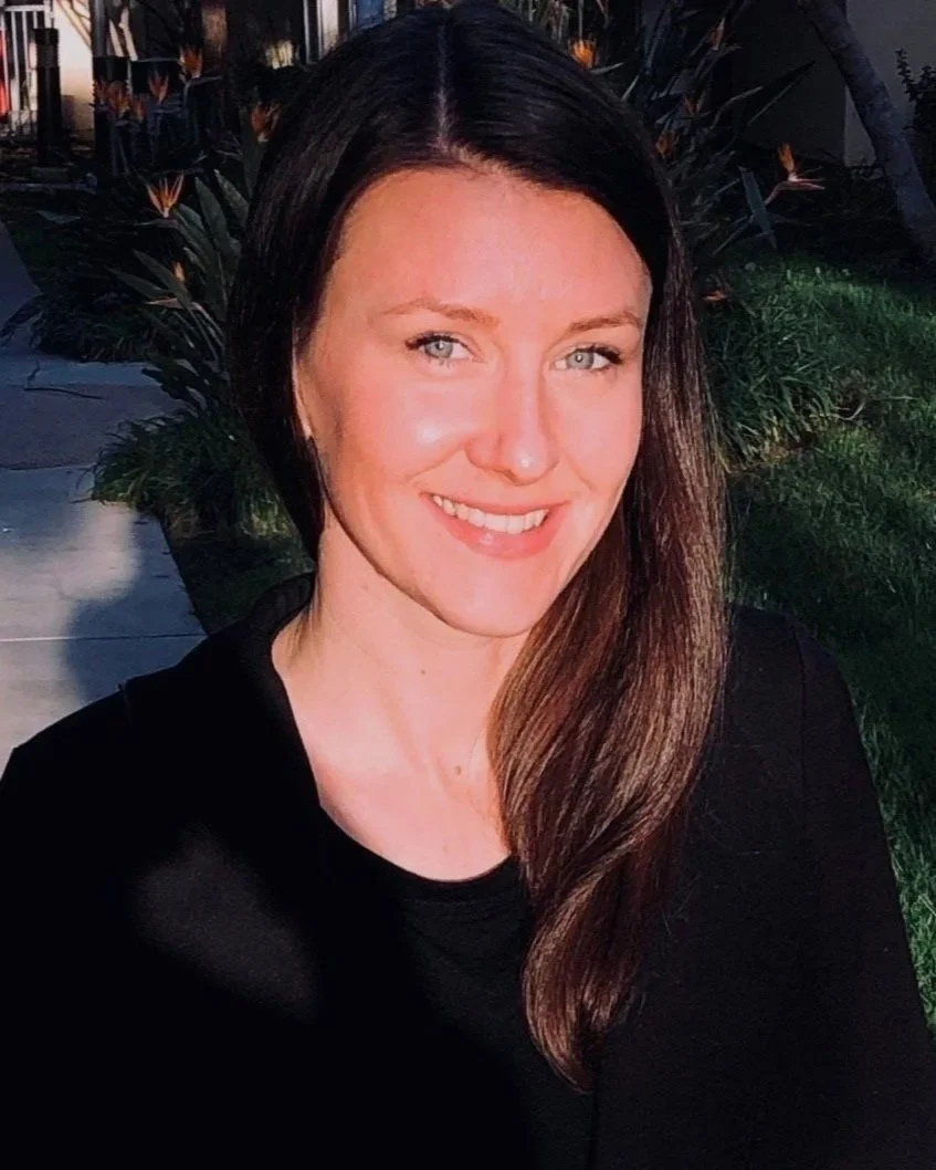 A woman with long brown hair and blue eyes smiling outdoors in front of green plants, wearing a black top.