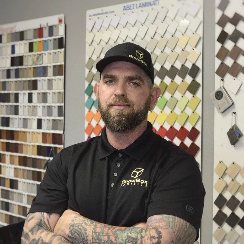 Showbox Exhibits Man with a beard and tattoos wearing a black polo shirt and cap, standing in front of wall displays with color samples.