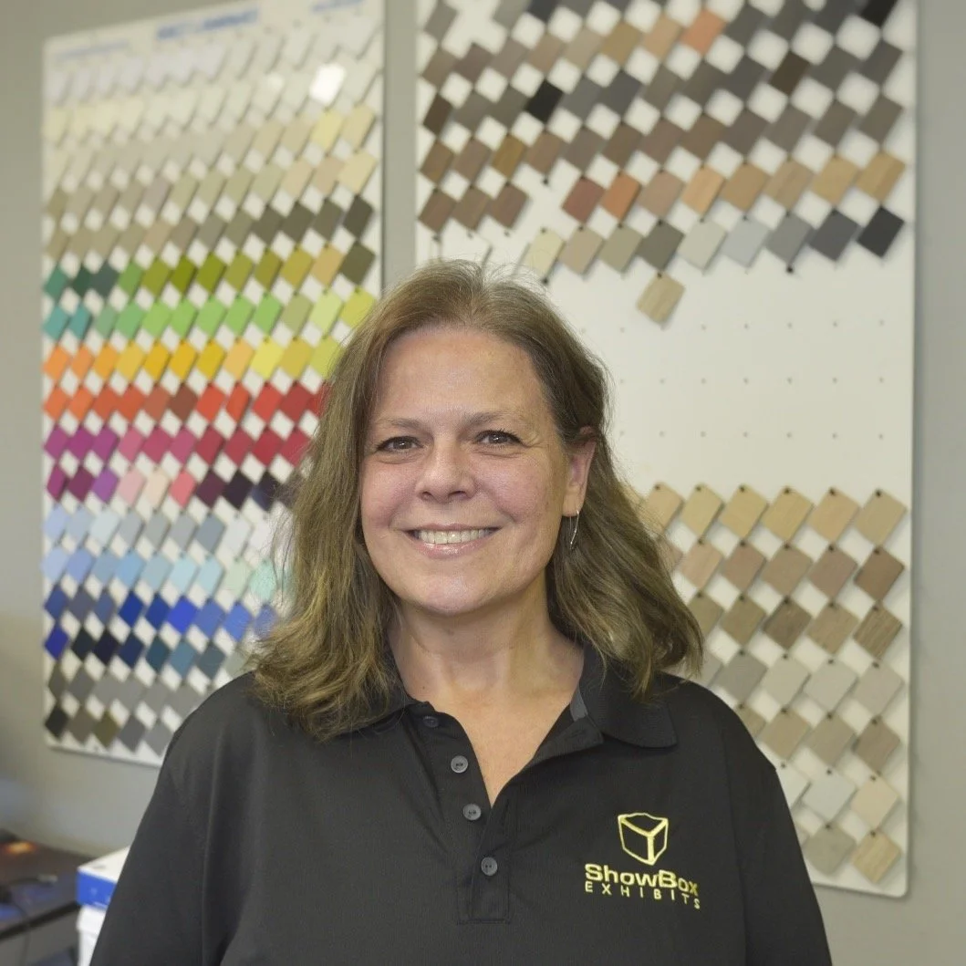 Woman smiling in front of color swatch panels, wearing a black ShowBox Exhibits polo shirt.