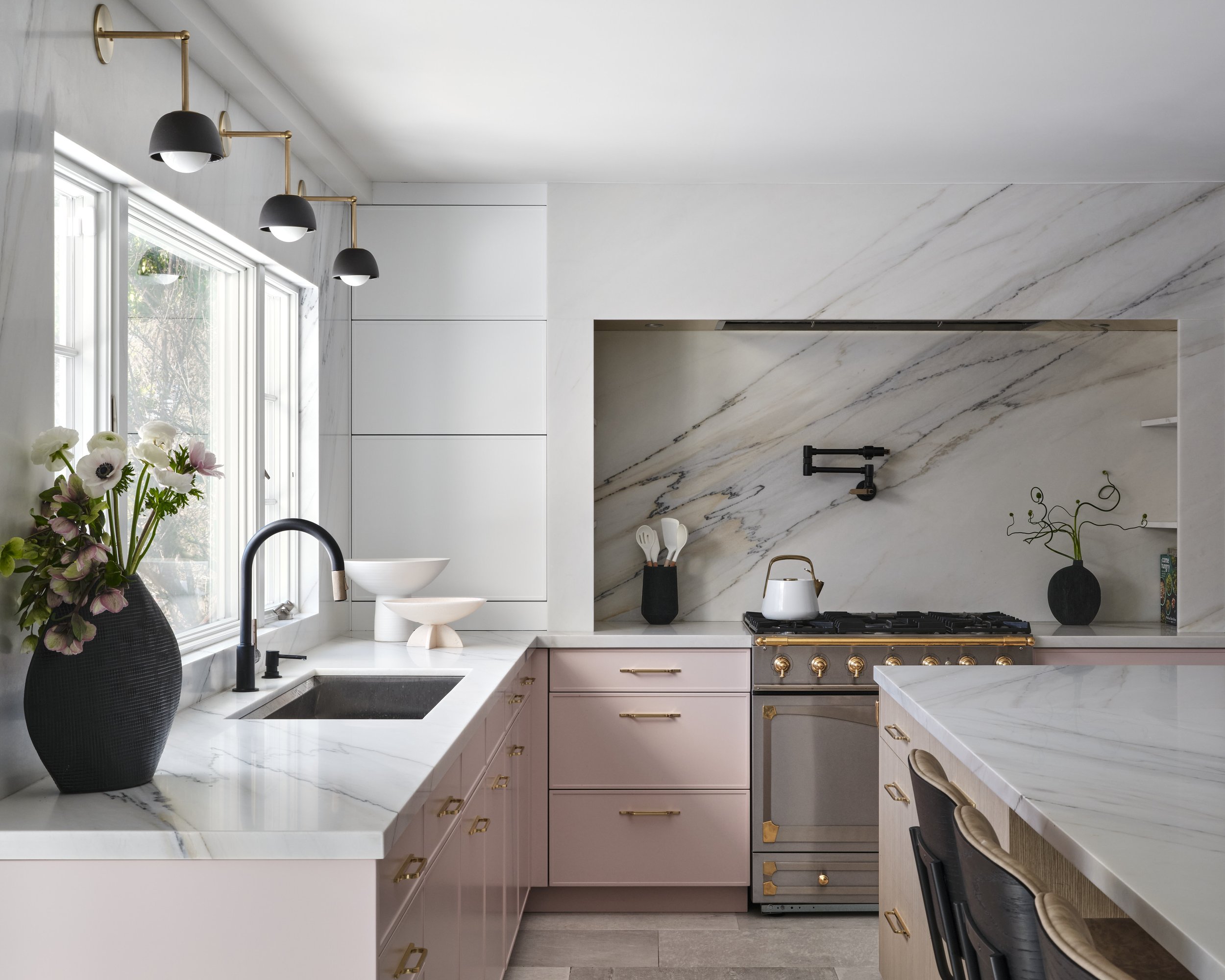 Modern luxury kitchen designed by Amy Oldis Design featuring custom pink cabinetry, white marble countertops, black and brass appliances, sculptural black pendant lighting, and curated floral accents.