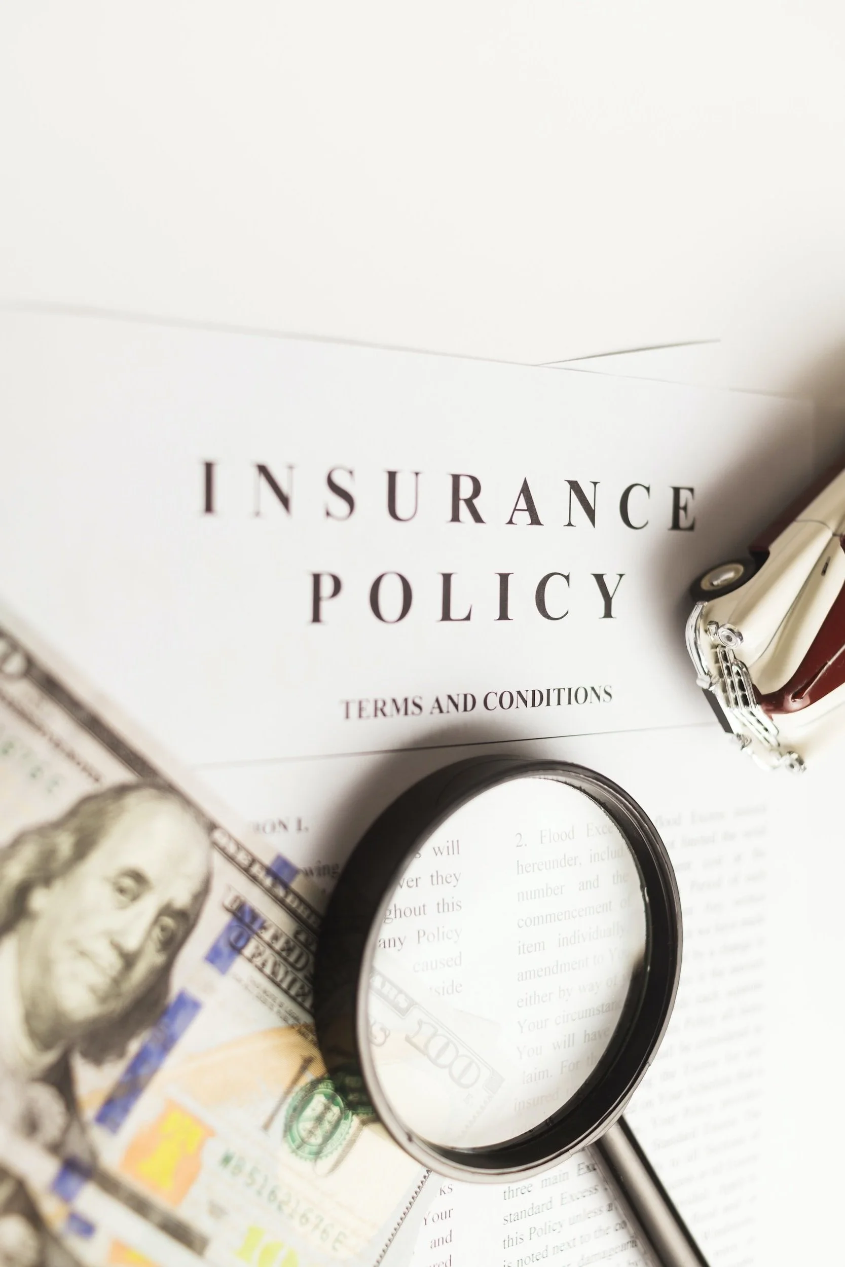 Insurance policy document with magnifying glass, hundred-dollar bill, and pen on white background.