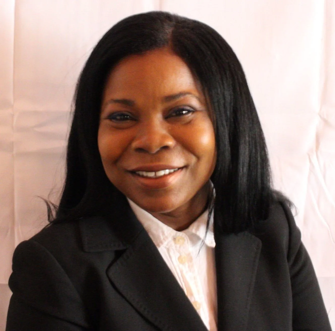 A woman with black hair wearing a black blazer and white shirt, smiling in front of a white background.