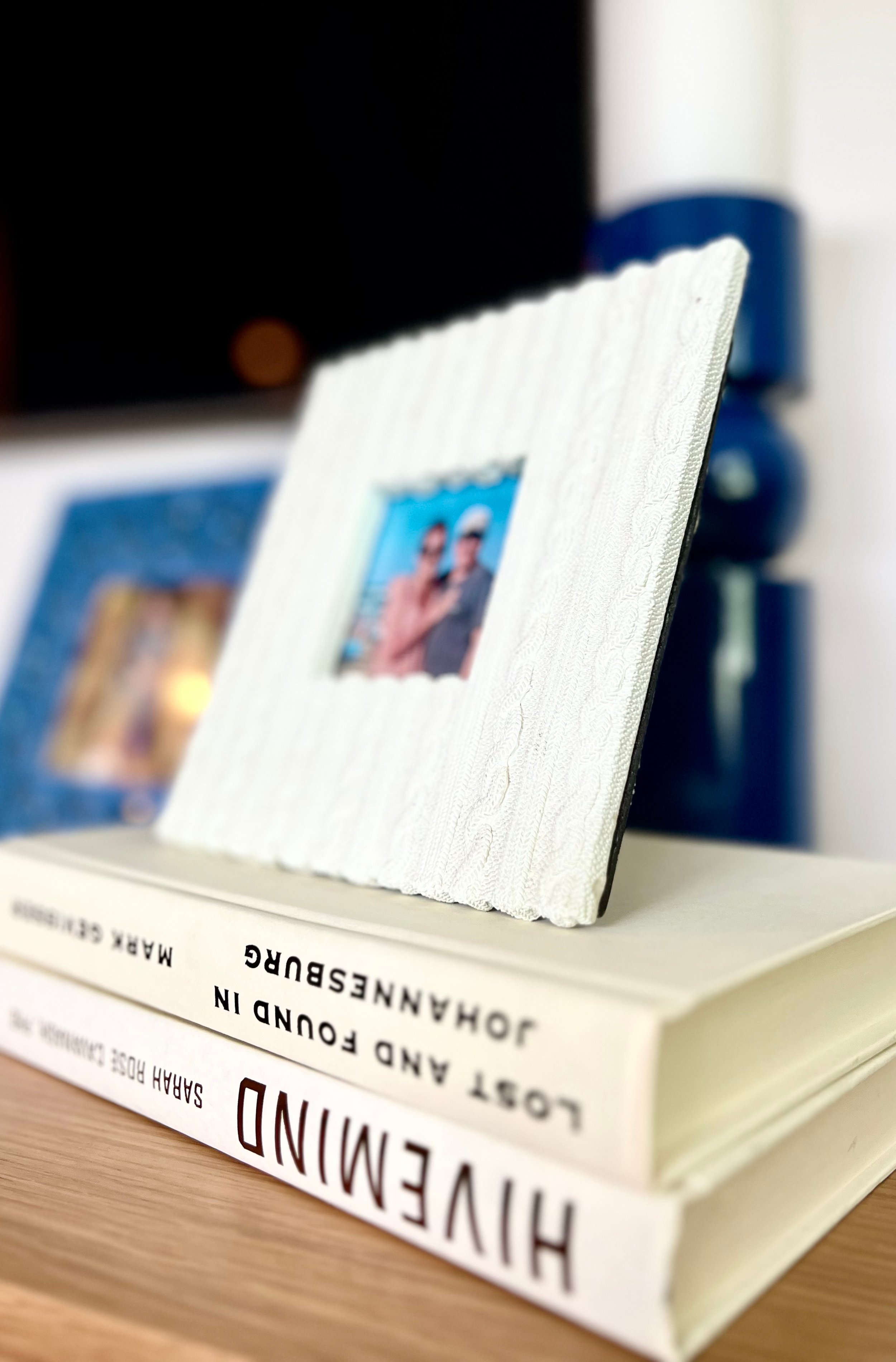 Close-up of a white book titled "HIVEMIND" by Sarah Doud, with a photo frame on top showing two people at the beach, and another photo frame blurred in the background, all placed on a wooden surface.