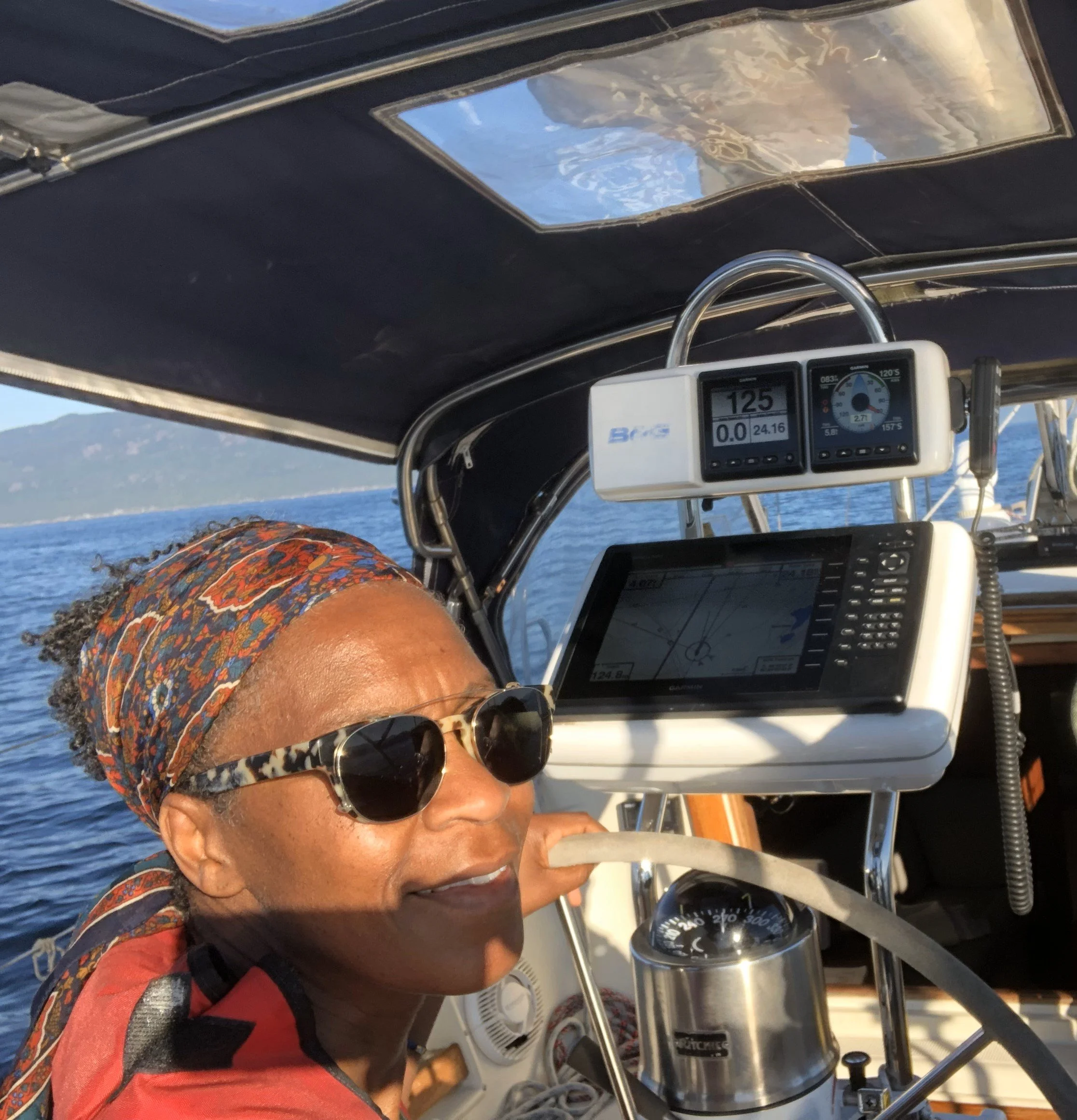Woman with sunglasses and headscarf smiling on a boat with navigational equipment and water in the background.