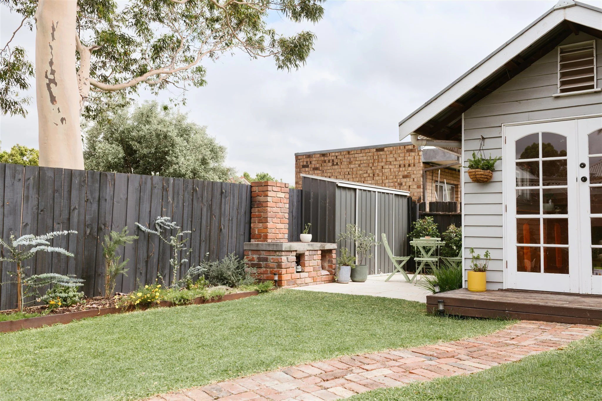 A backyard garden with a brick pathway, a wooden deck, and a white house with glass-paneled doors. There is a black wooden fence, potted plants, a small table with chairs, and a hanging plant basket.