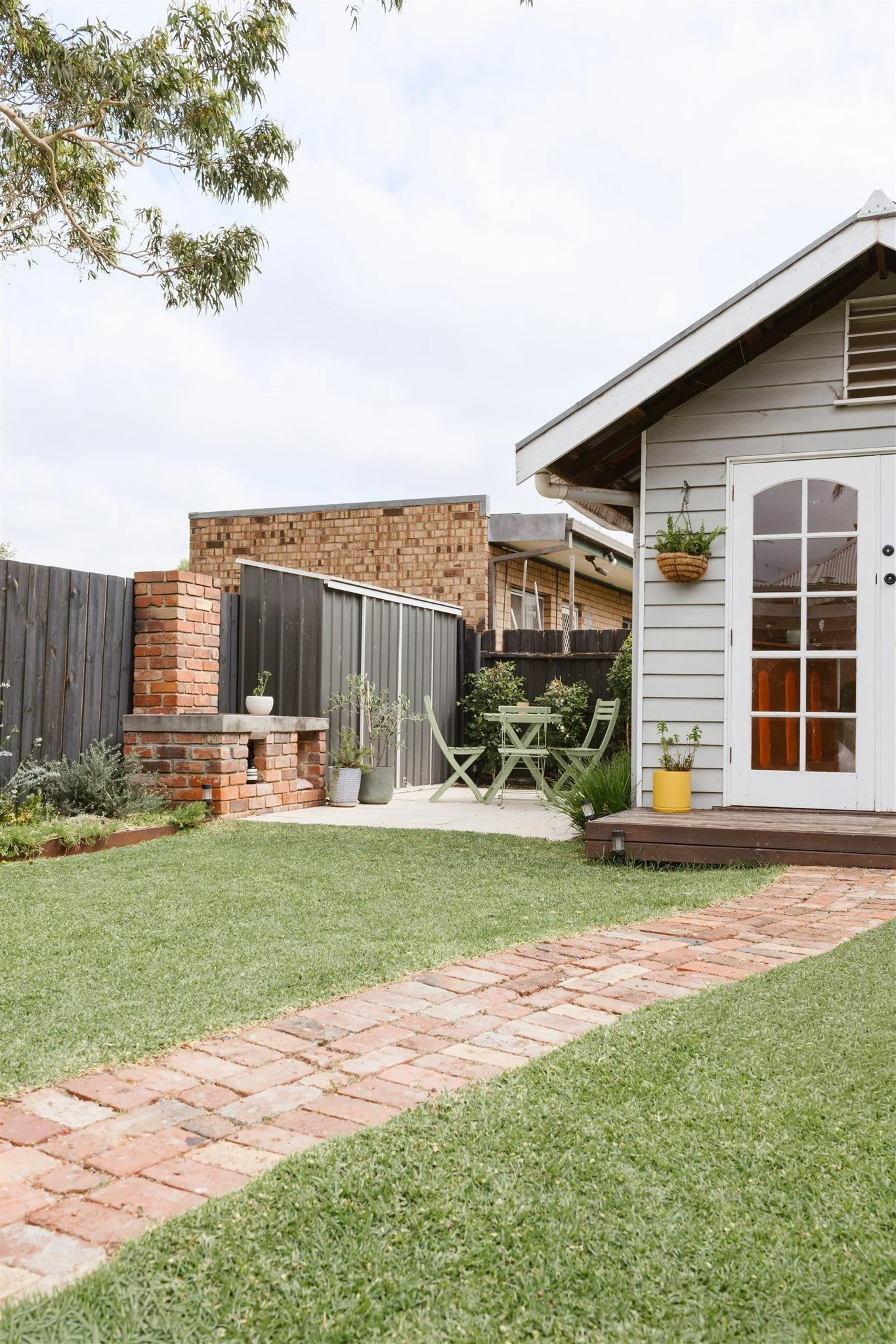 A backyard with a grassy lawn, brick pathway, small patio with green outdoor dining set, a white shed with a glass door, potted plants, and a brick wall with a decorative brick structure.