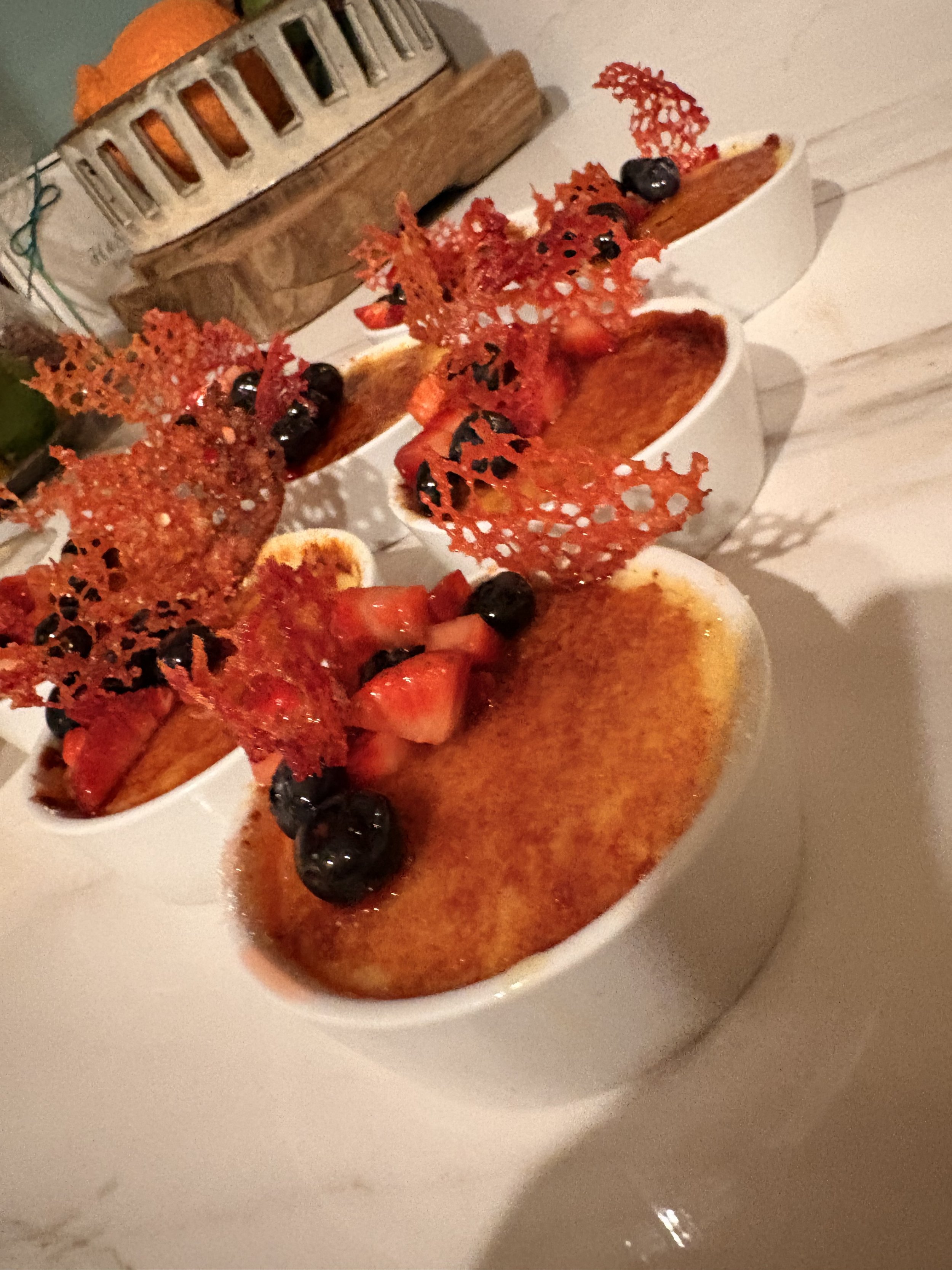 Three ramekins of crème brûlée topped with fresh strawberries, blueberries, and decorative caramelized sugar lace on a white table.
