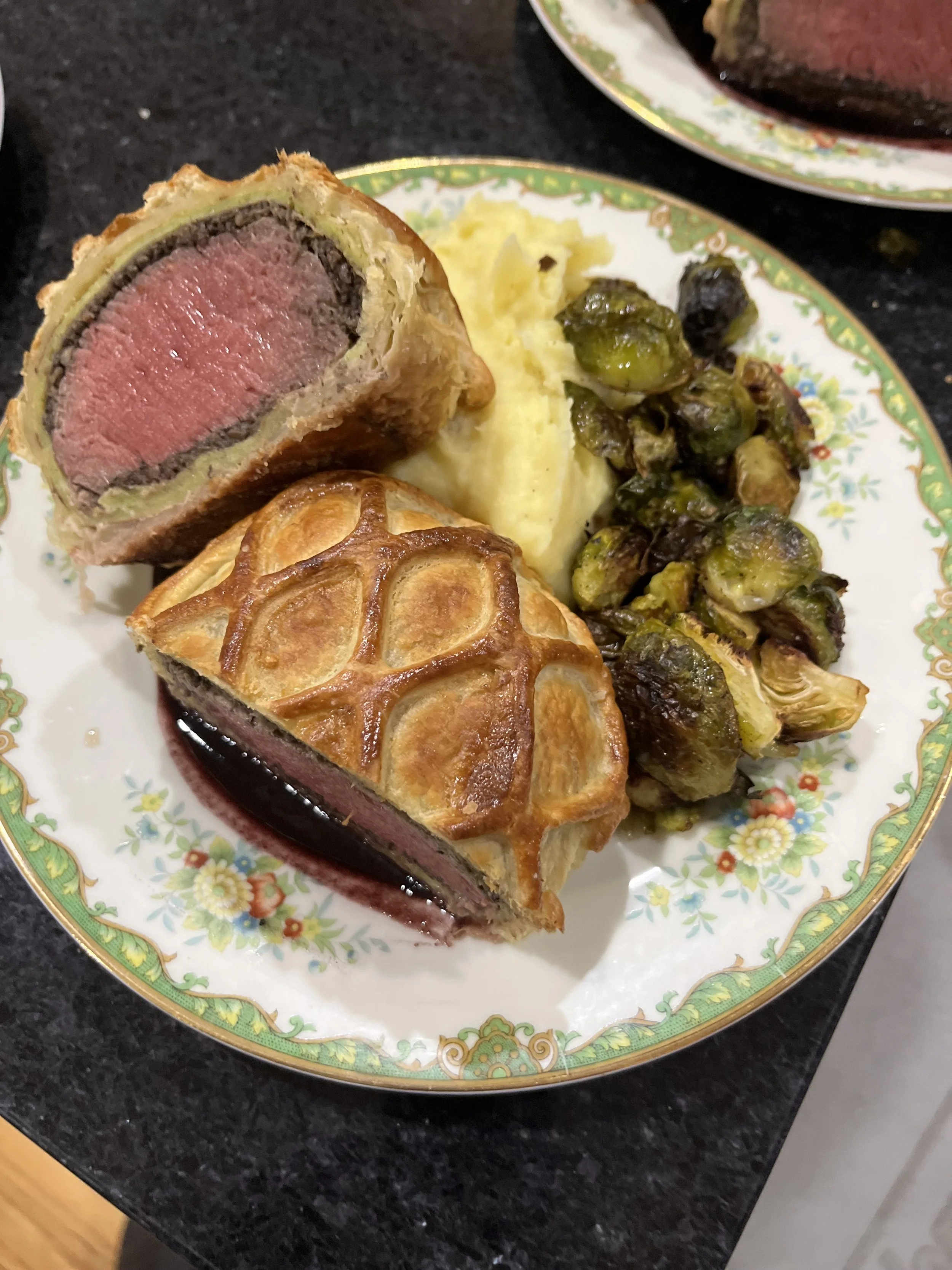 A plate with two slices of beef Wellington, mashed potatoes, and roasted Brussels sprouts.