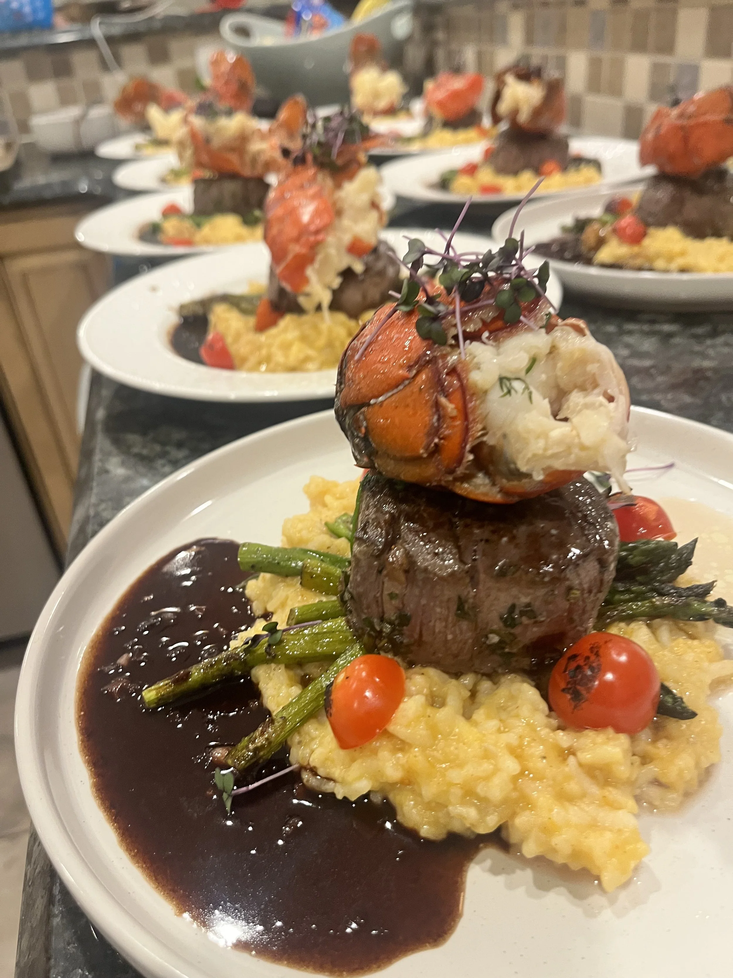 Plate with filet mignon topped with grilled lobster, garnished with microgreens, served with green beans, cherry tomatoes, mashed potatoes, and a dark sauce.