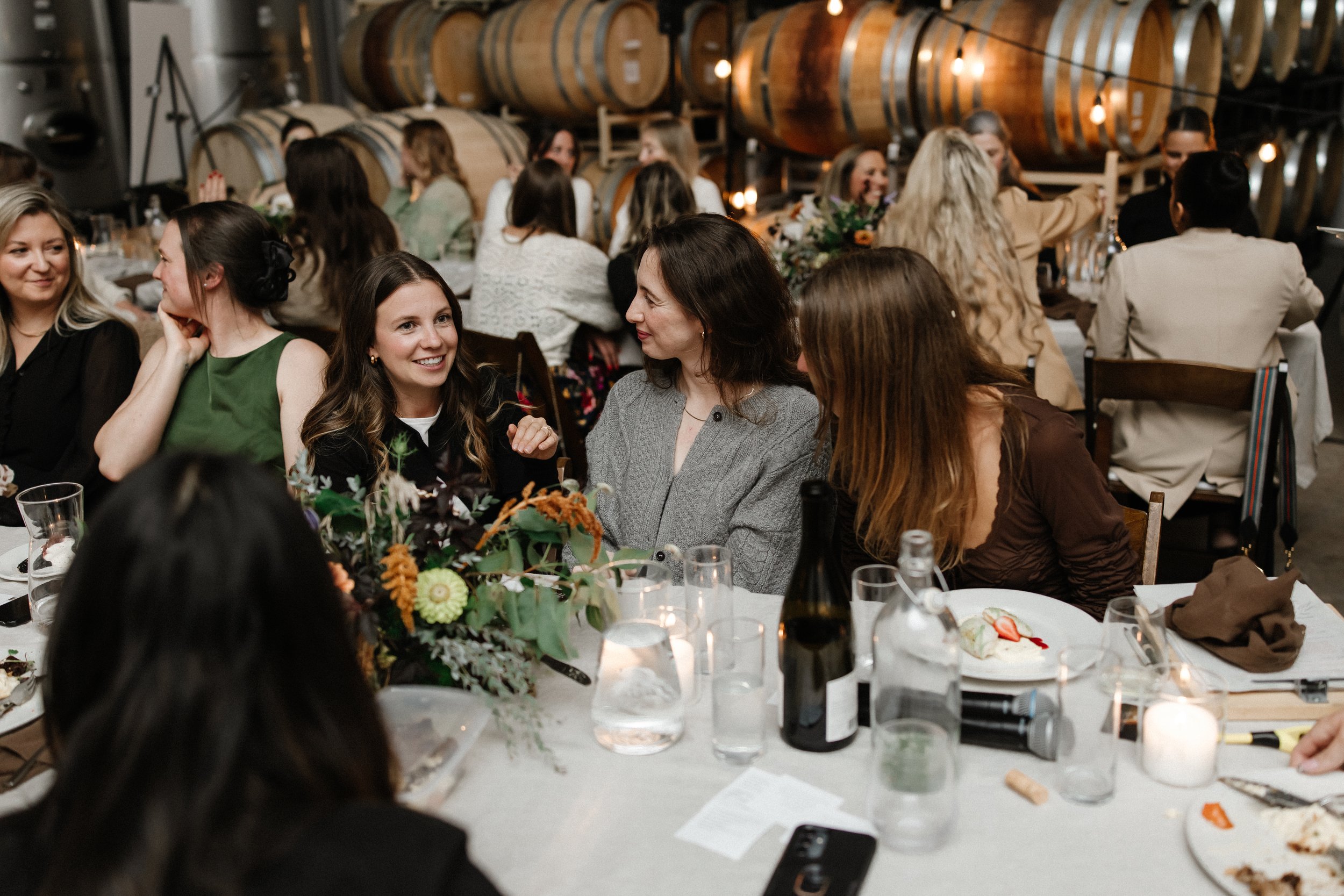 Women sitting at a dinner table, engaging in conversation, with wine bottles, glasses, floral centerpieces, and desserts in a rustic event space with large wine barrels in the background.
